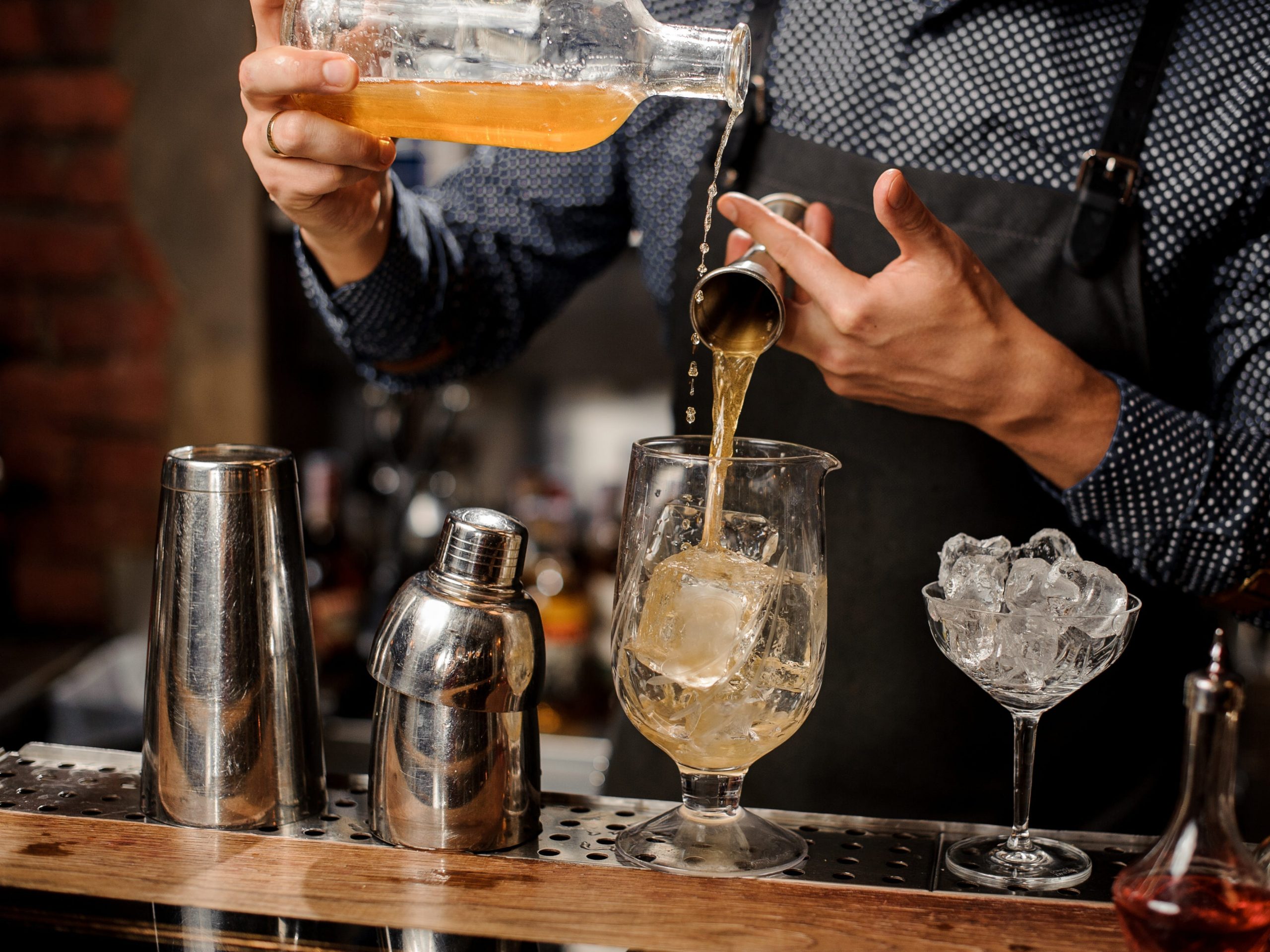 Bartender in black apron and blue shirt mixing whiskey, syrup and ice in cocktail glass at a bar counter