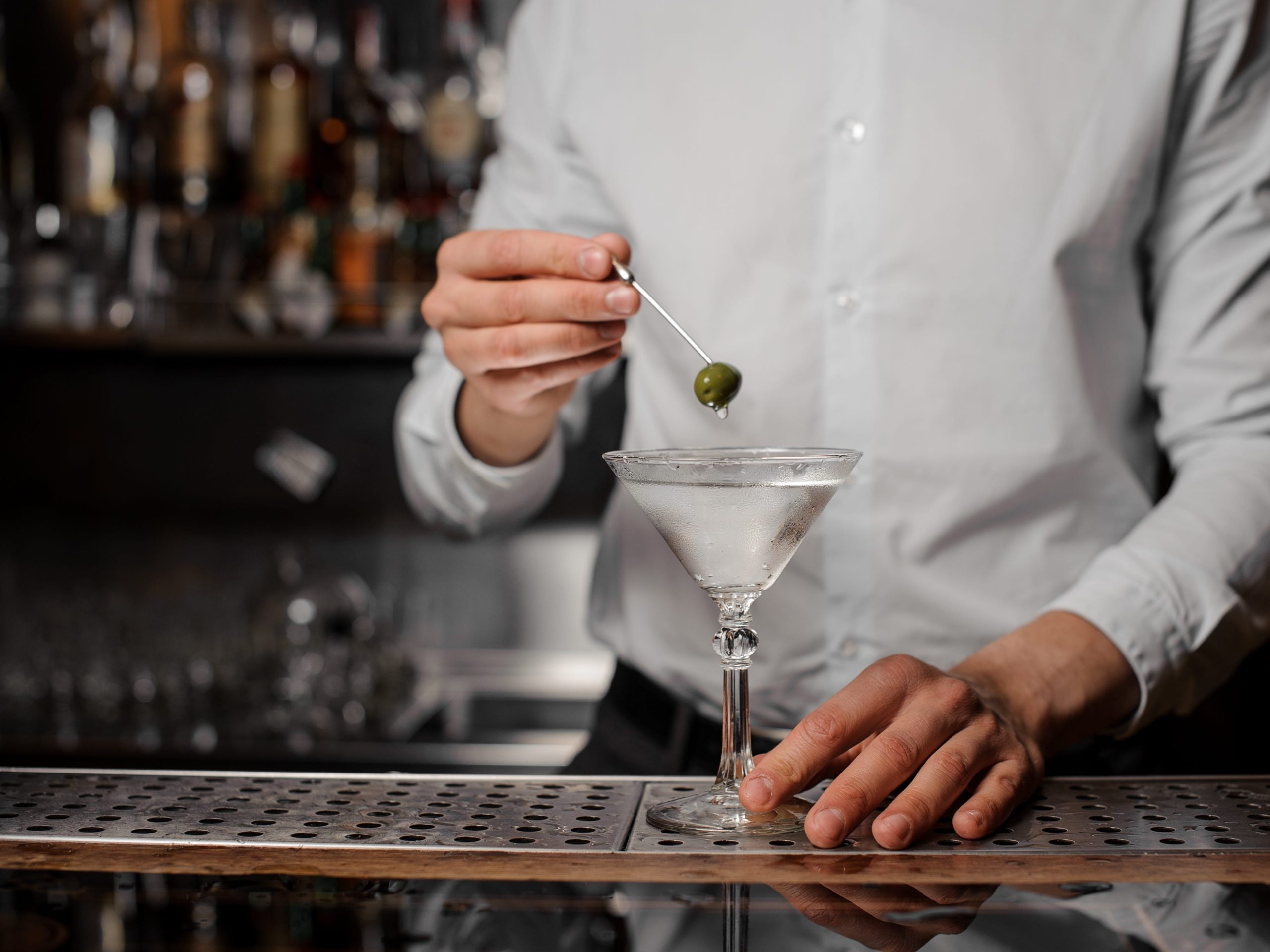 Bartender adding an olive on the skewer into the martini glass with transparent alcoholic drink on the bar counter