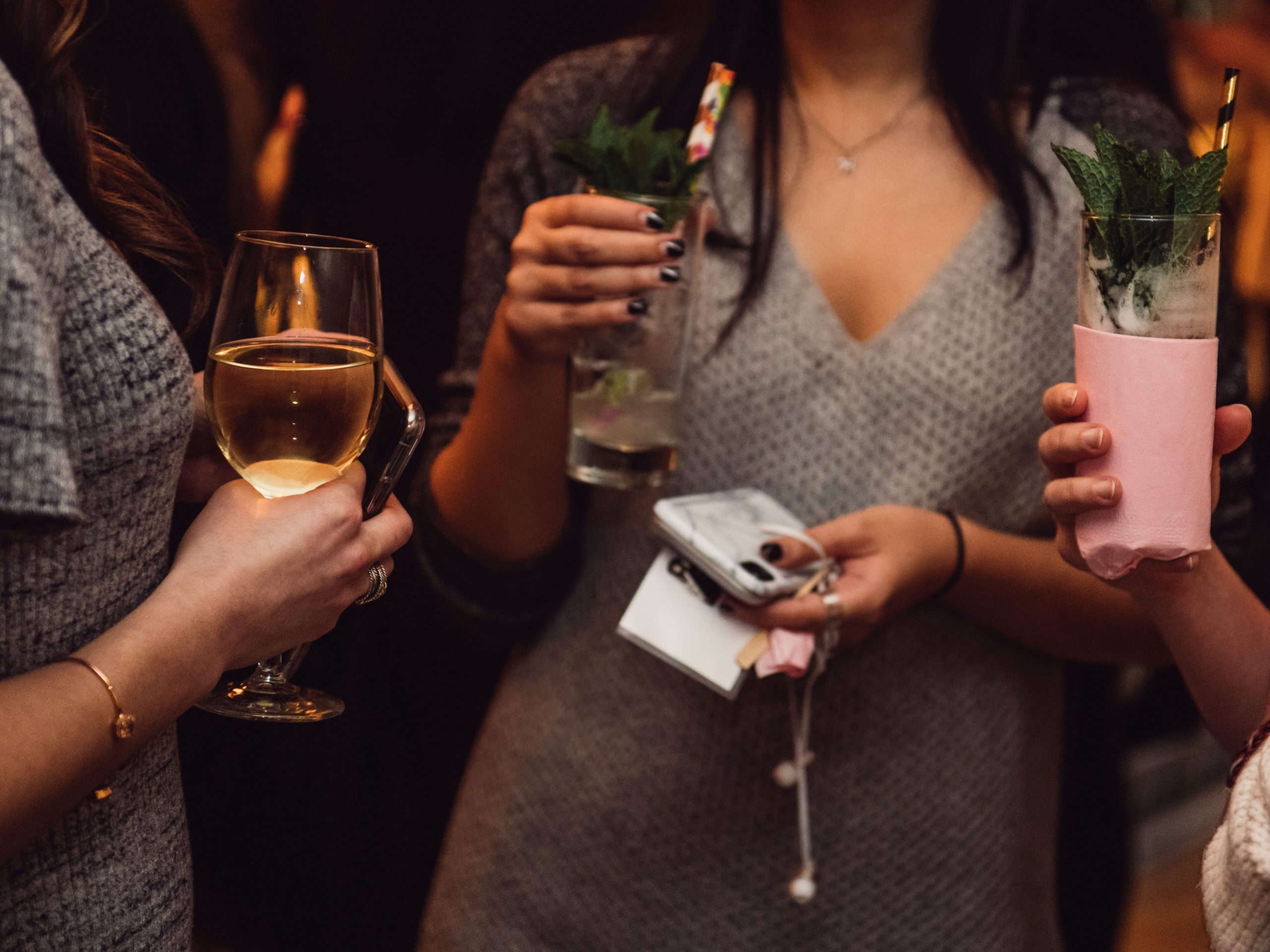 Women drinking cocktails and wine and talking with guests at a bar.