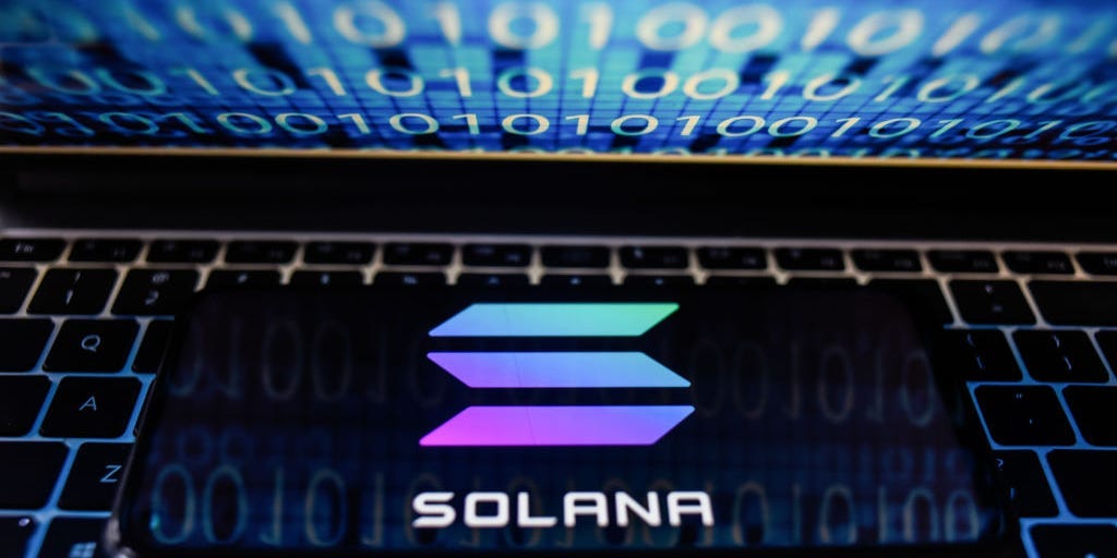 In this photo illustration a Solana logo seen displayed on a smartphone with binary code on a laptop screen