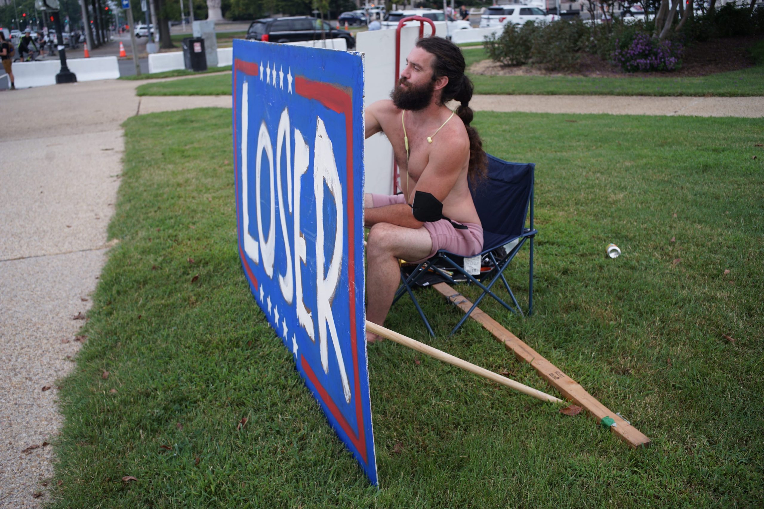 A shirtless man sits with a large sign that says Loser.
