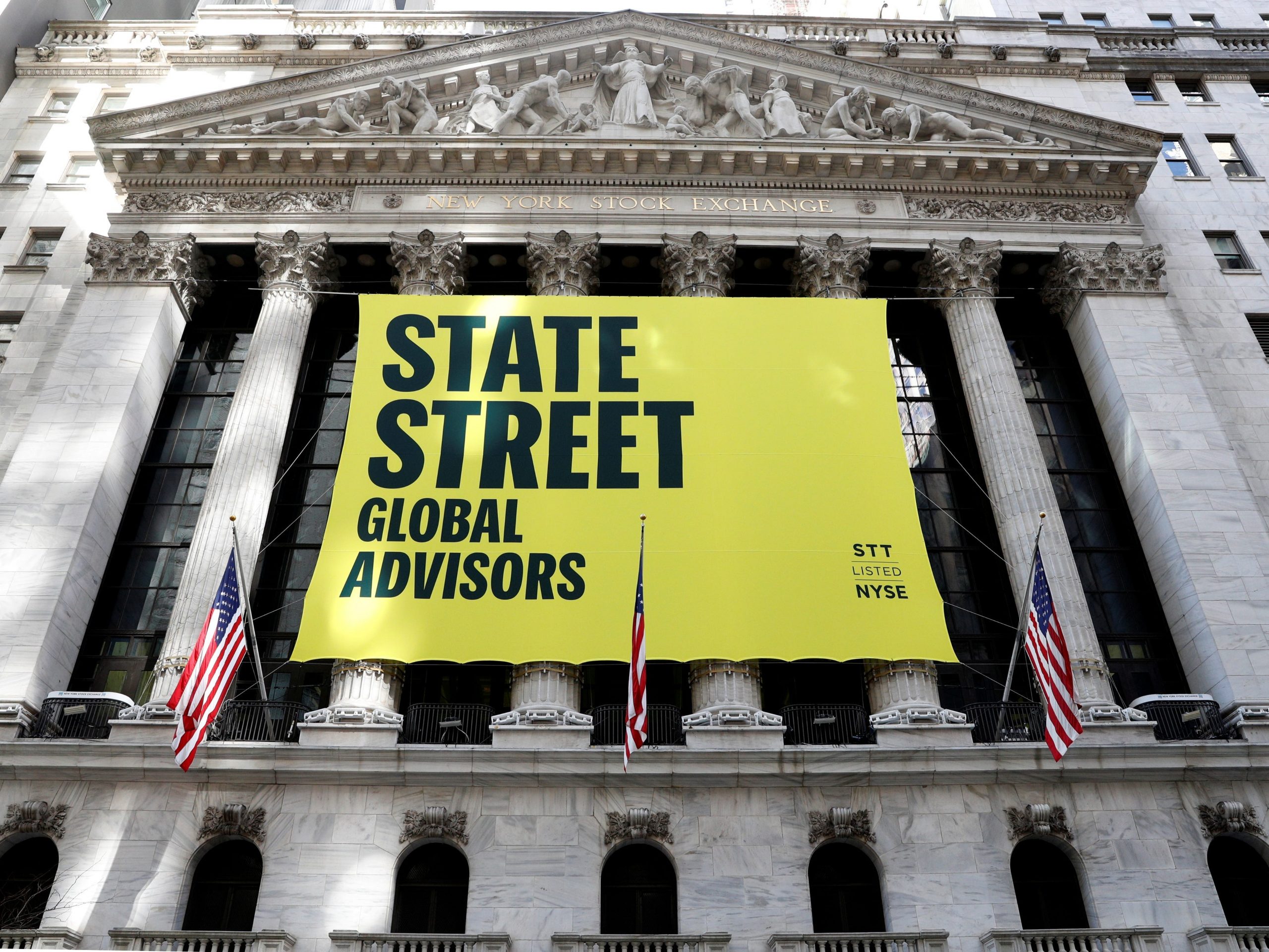 A large yellow sign that reads "State Street Global Advisors" hangs on beige-colored pillars in front of the New York Stock Exchange. American flags hang in front of the large yellow sign.