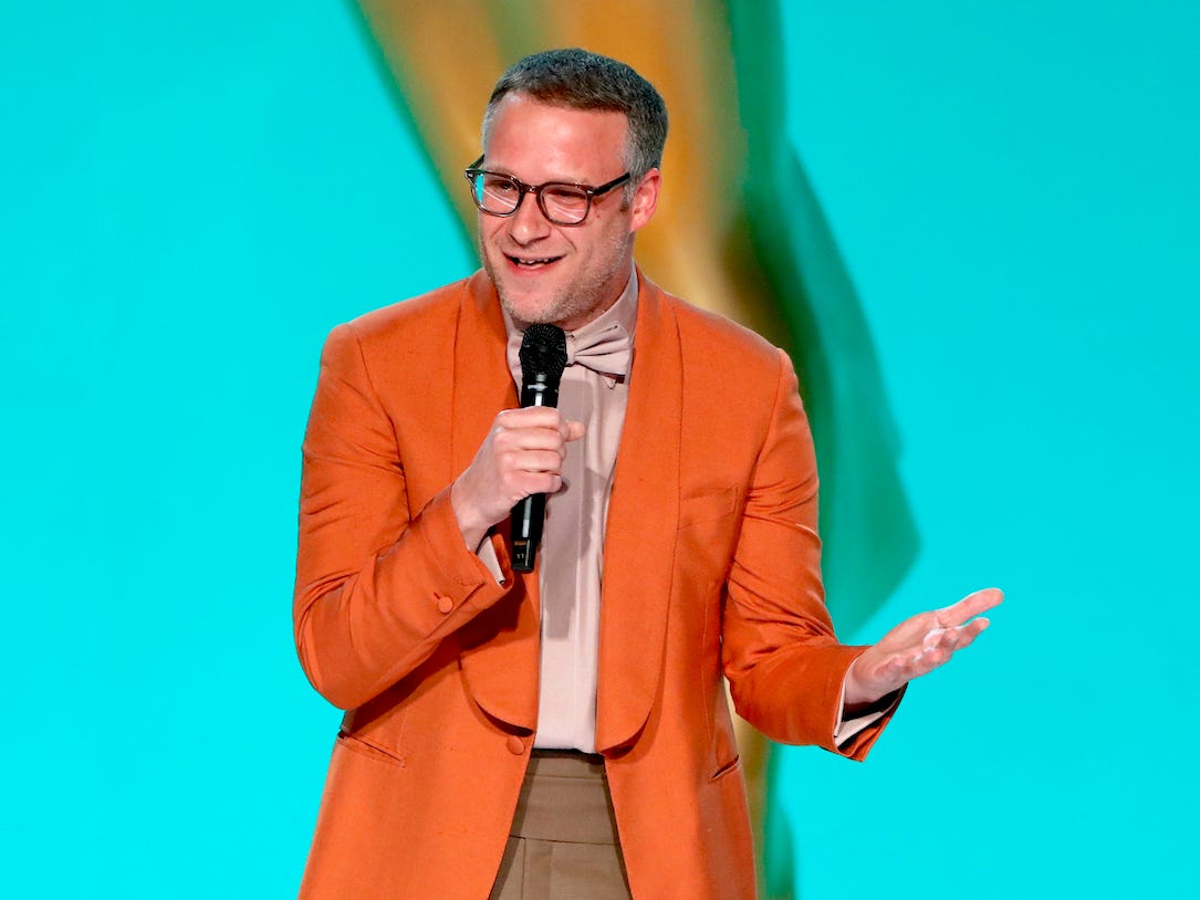 Seth Rogen (wearing an orange blazer and brown pants) standing in front of a blue backdrop.