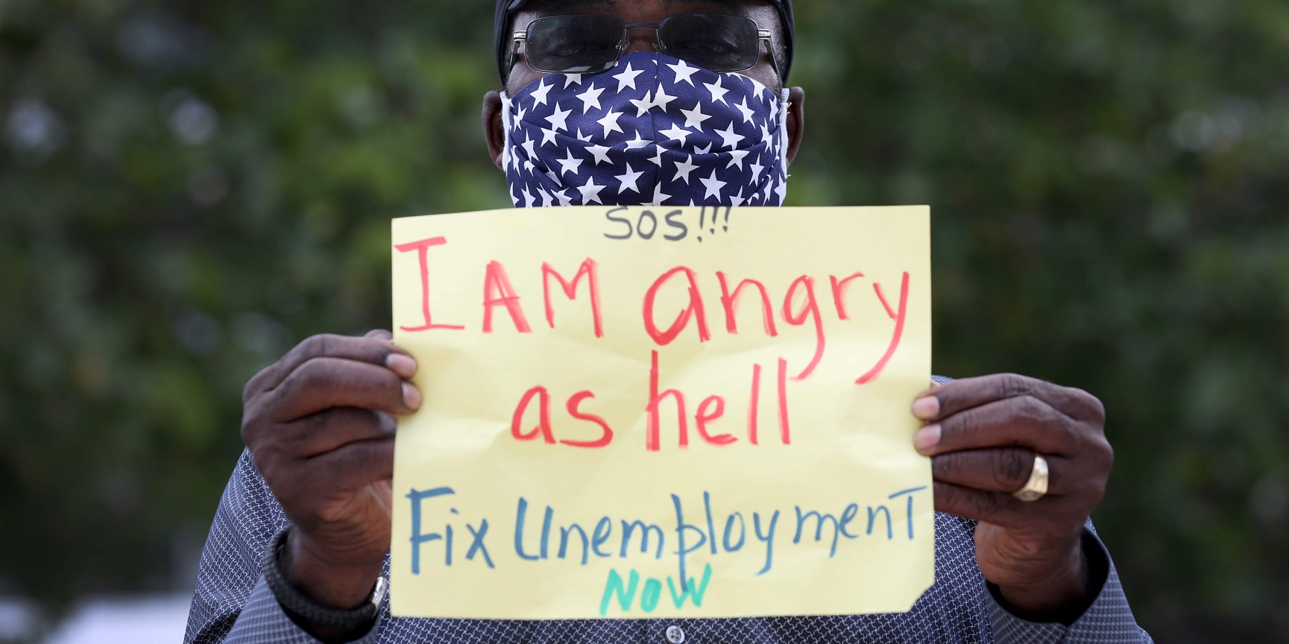 an unemployed worker holds a sign that says  I Am angry as hell Fix Unemployment Now,'