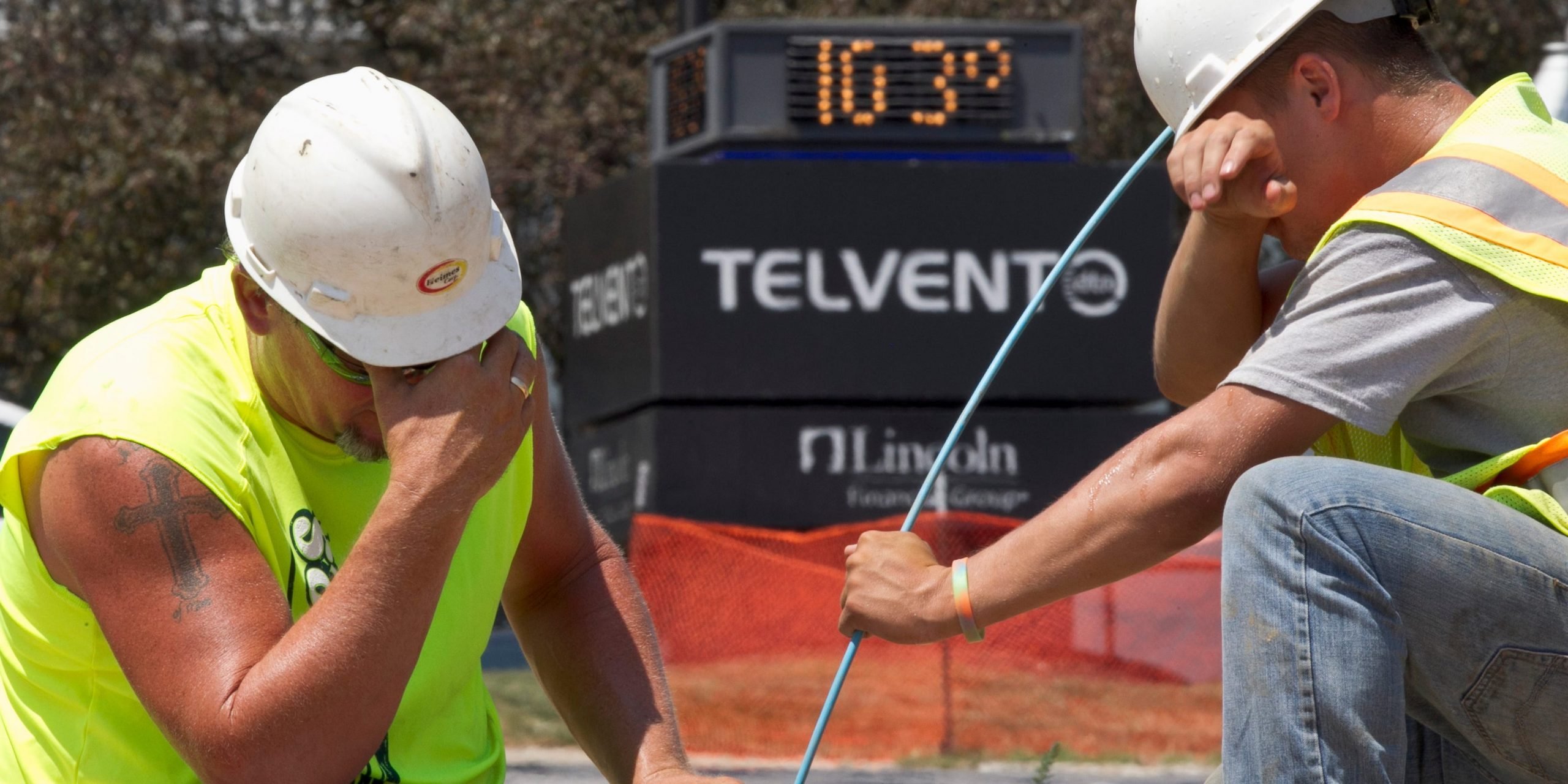 Construction workers wipe faces in front of digital sign showing temperature is 103 degrees.