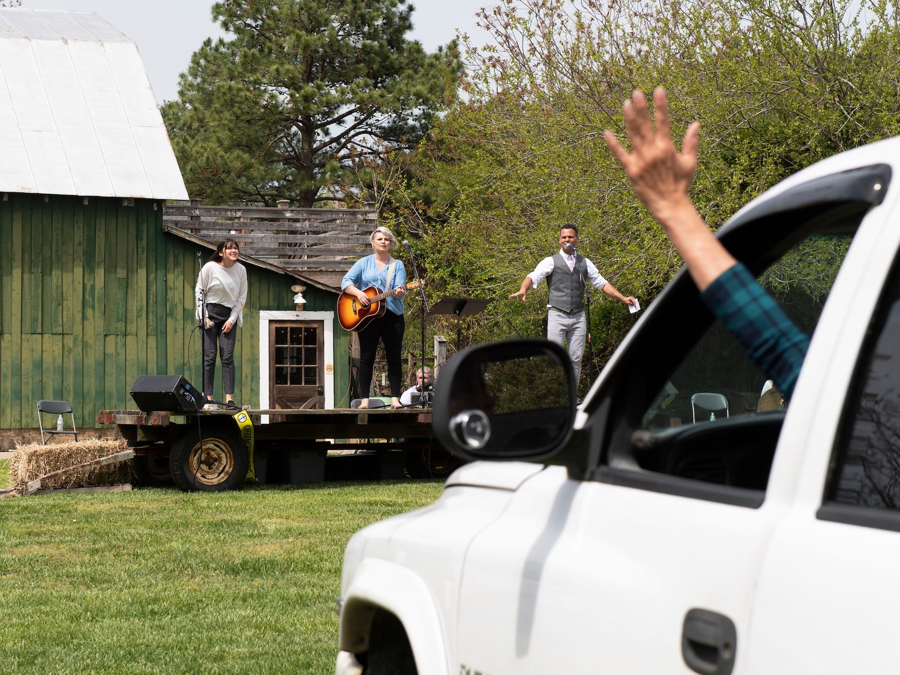 The arm of someone wearing a plaid shirt is stretched out of a white car parked on a lawn as if in celebration. In the background are three singers singer on an open air church in front of a green wood church.