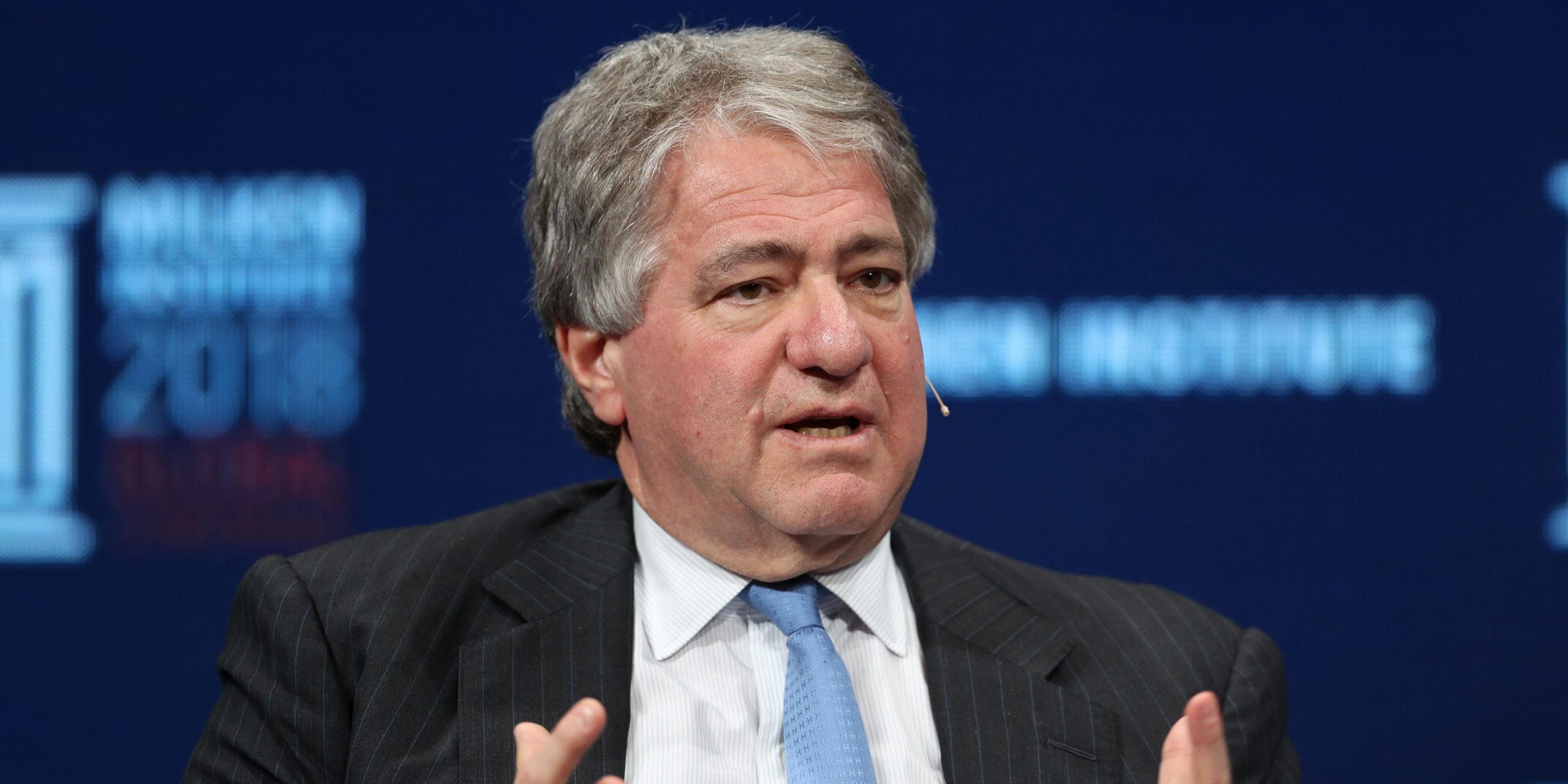 Leon Black wears a dark grey suit, white shirt, and blue tie while speaking on stage.