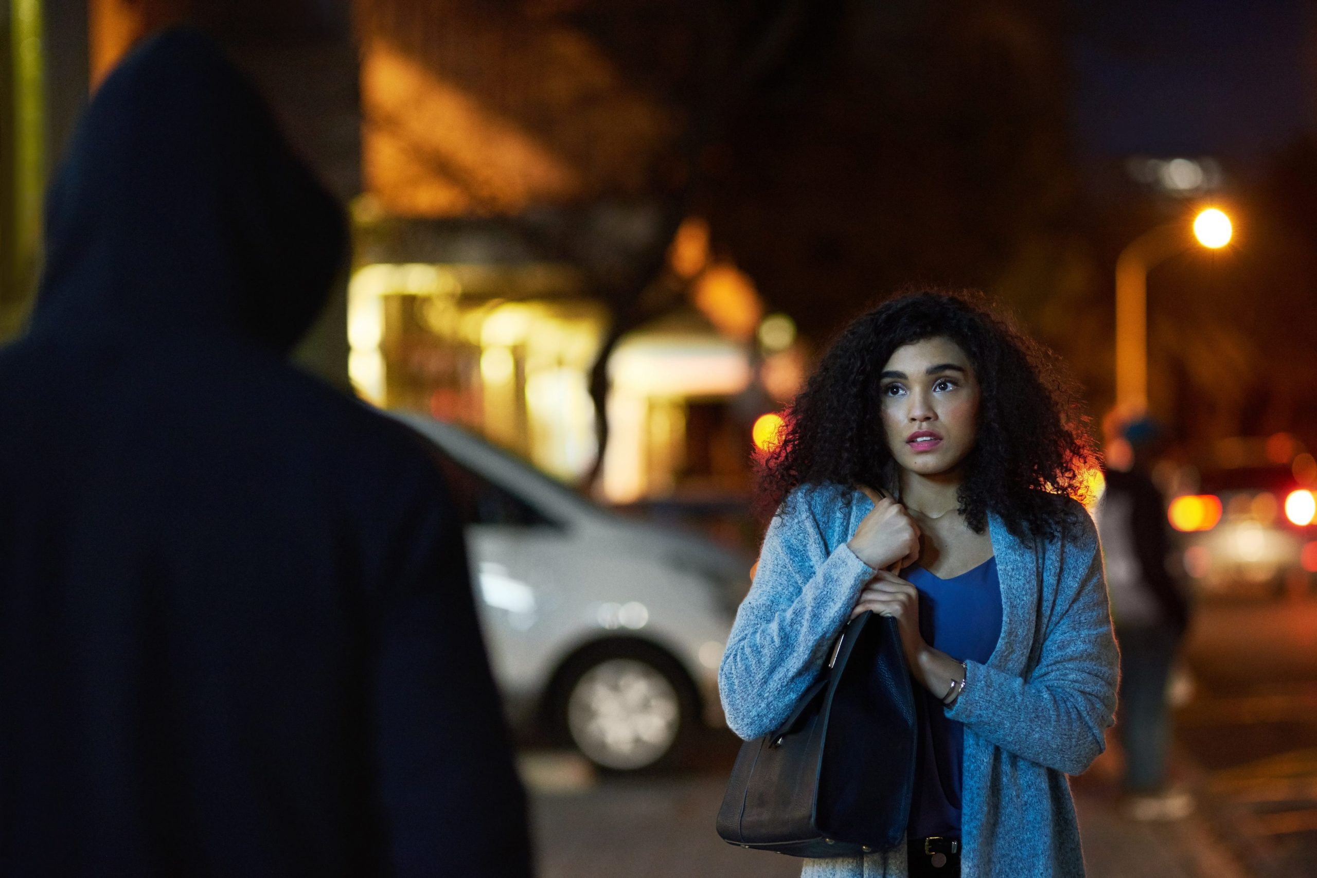 Young, Latina woman clutches purse as she is approached by a stranger in shadow.