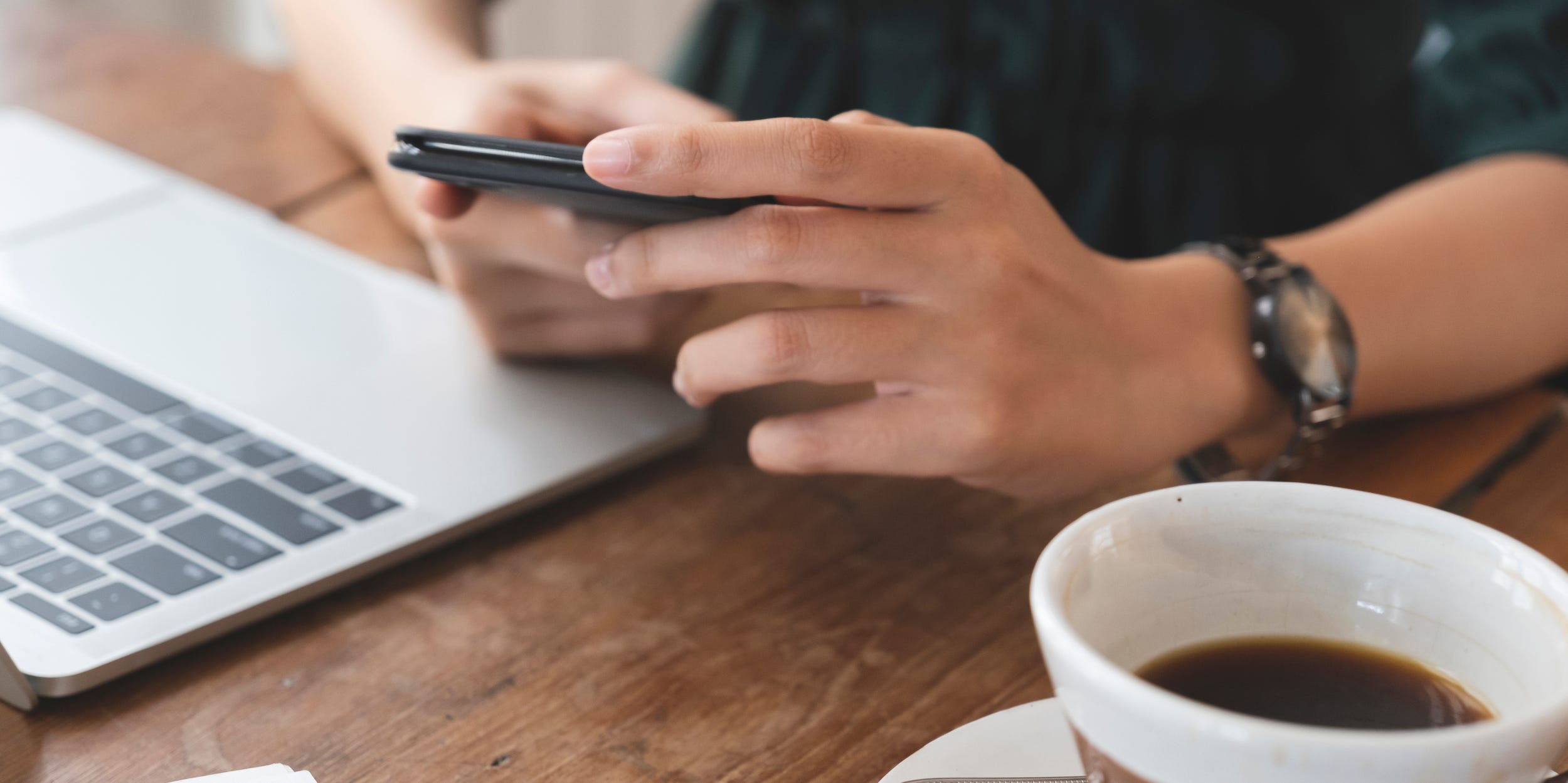 person at desk using phone and laptop with coffee cup