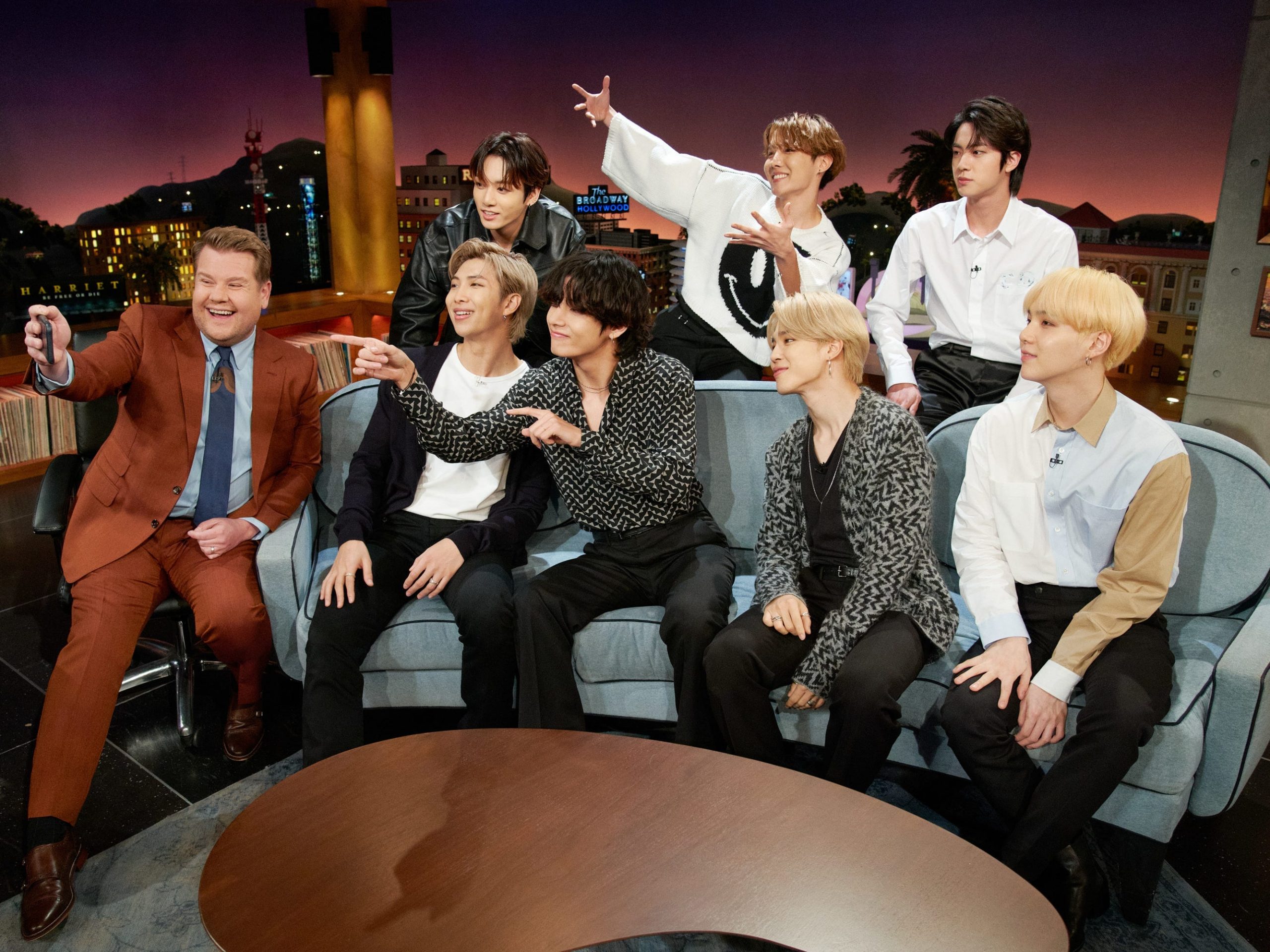 james corden on the set of the late late show taking a selfie with the seven members of bts, sitting on and standing behind a couch