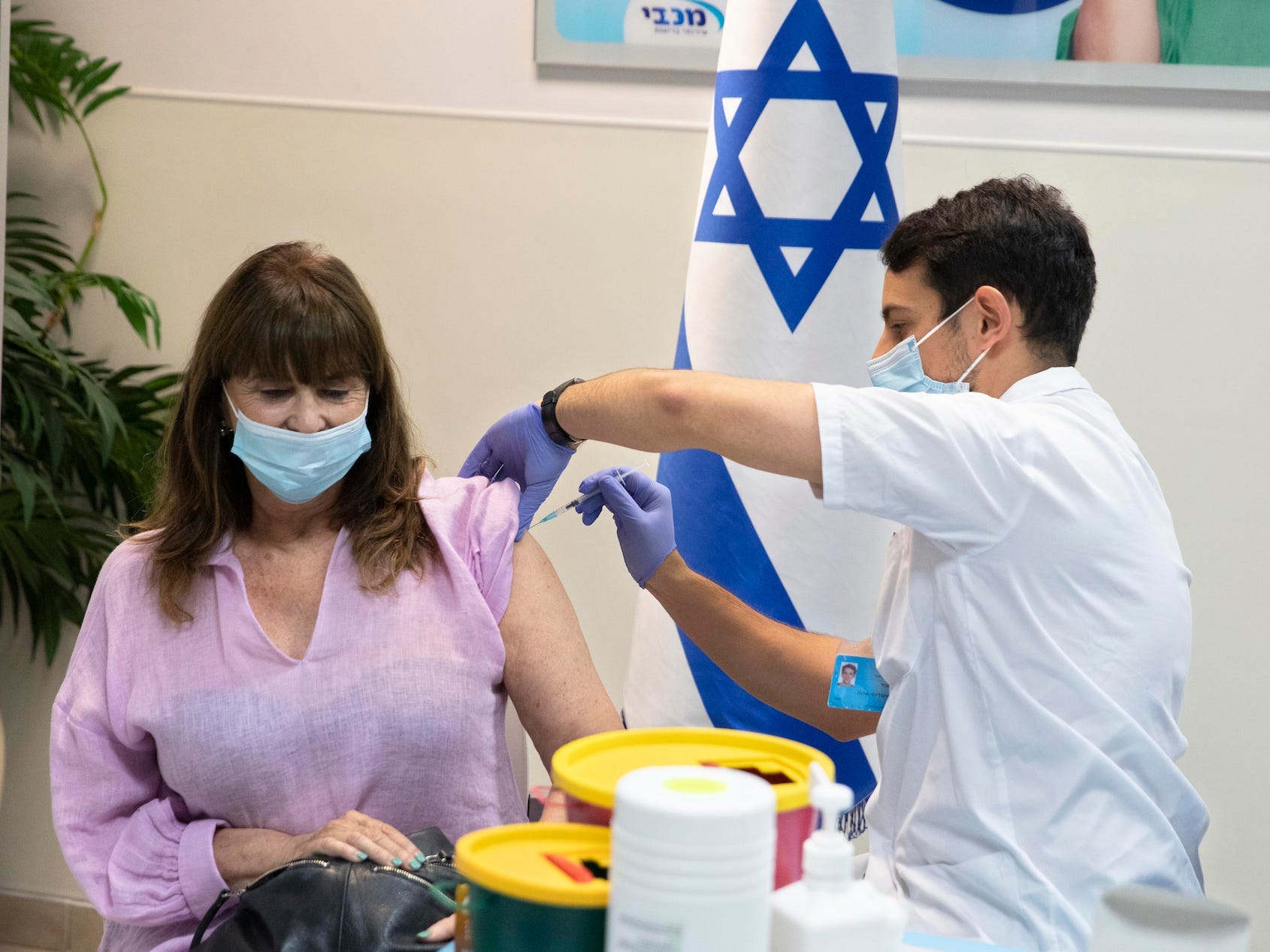 A masked woman wearing a purple shirt is getting vaccinated by a man in scrubs wearing a mask in front of an Israeli flag