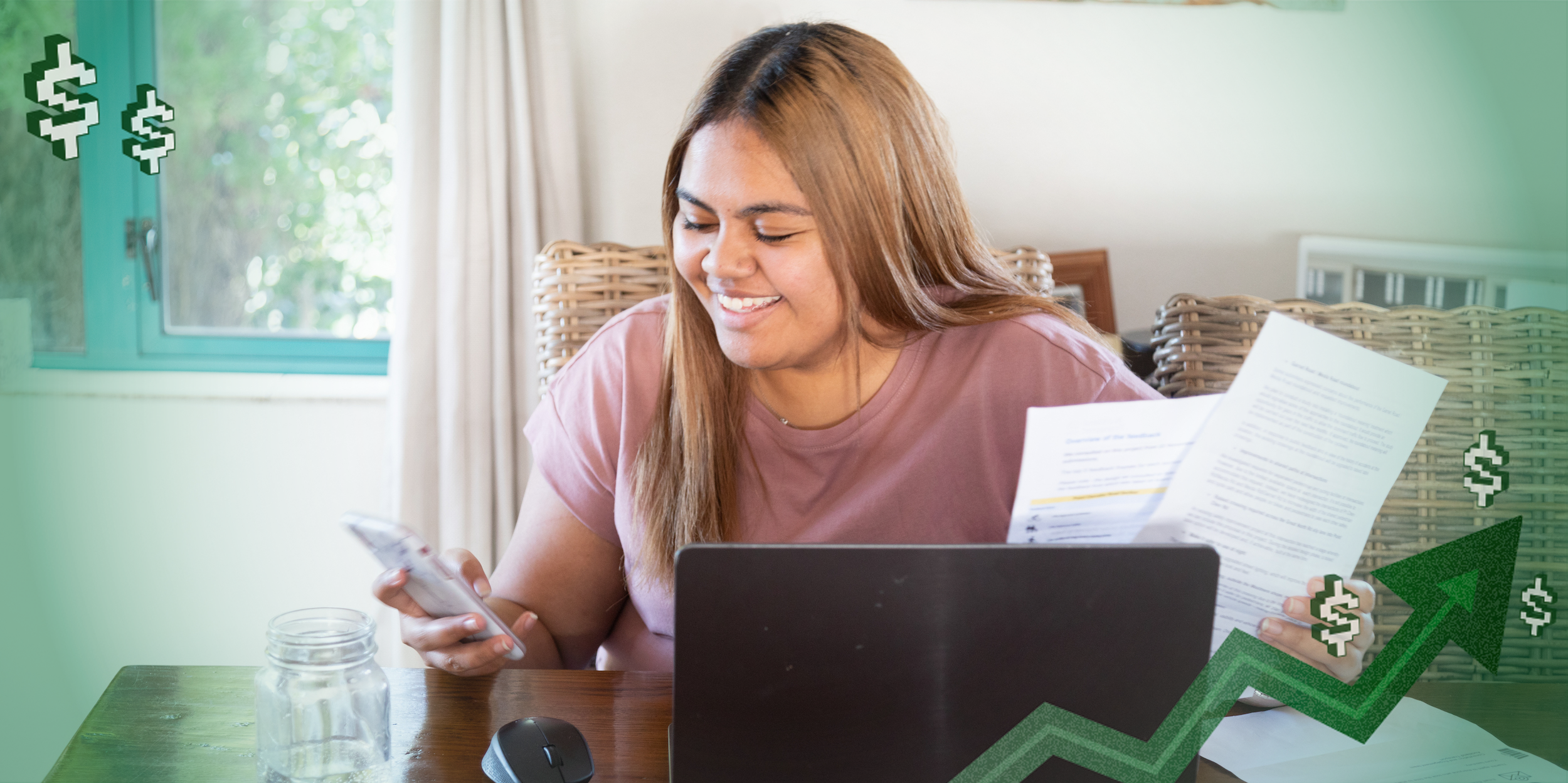 A young woman looking at her laptop and paperwork at a kitchen table.