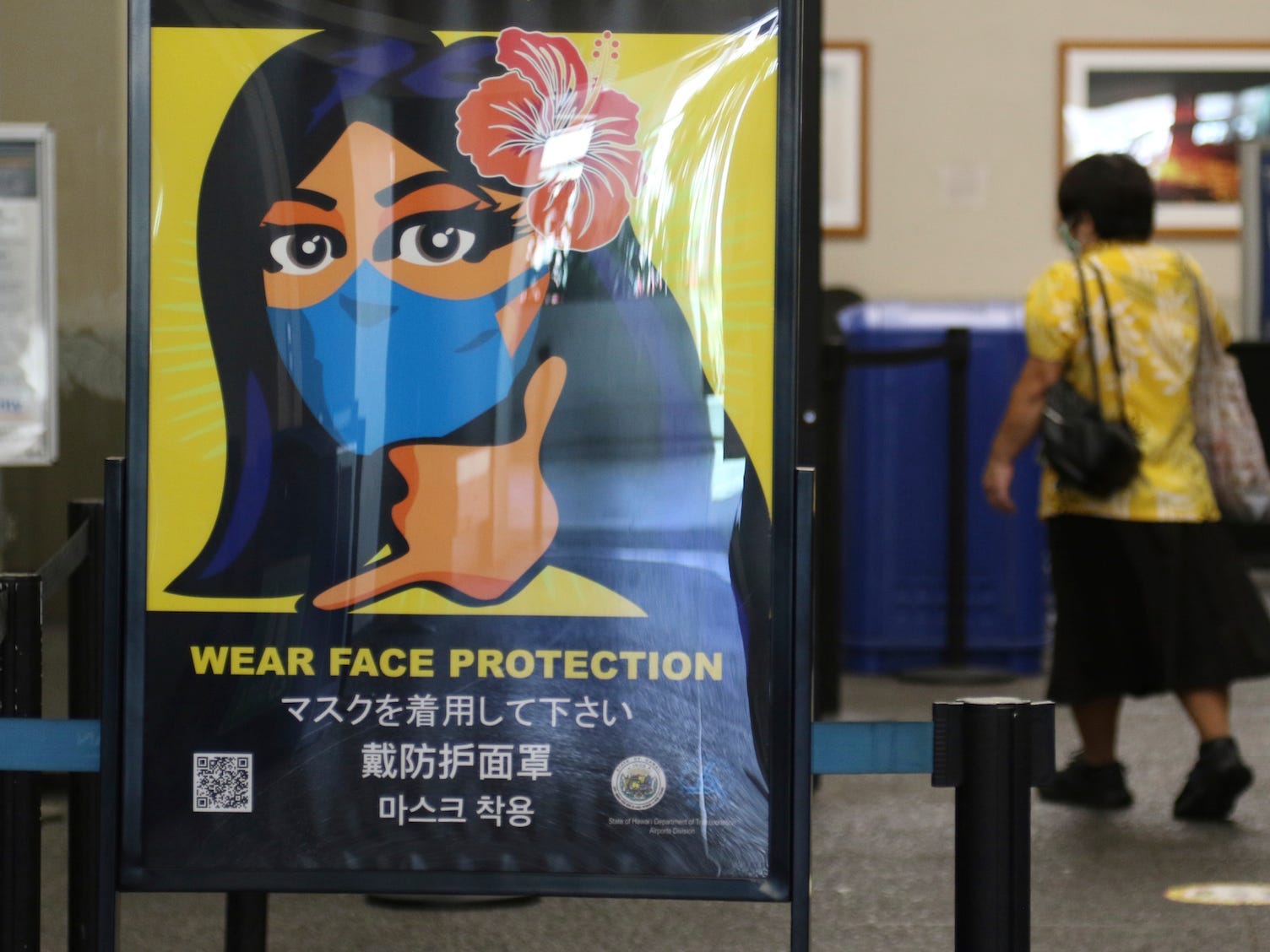 A poster shows a cartoon of a woman with a hibiscus flower in her hair wearing a blue mask, reading: "wear face protection."