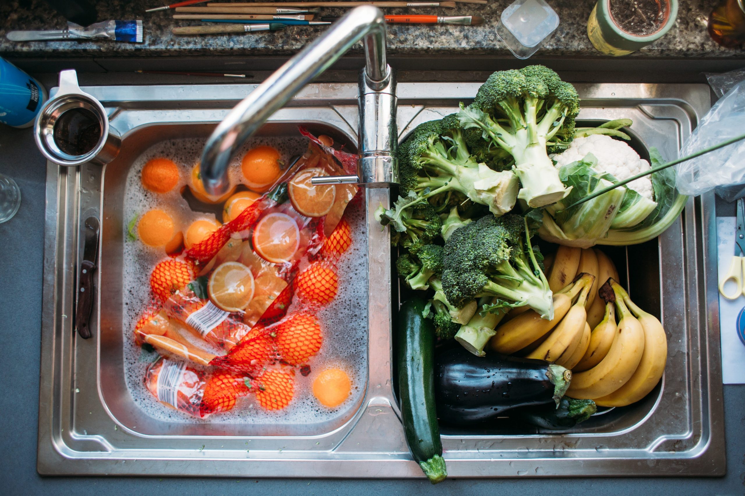 A double bowl kitchen sink with oranges floating in water in the left side and broccoli, eggplants, bananas resting in the right side.