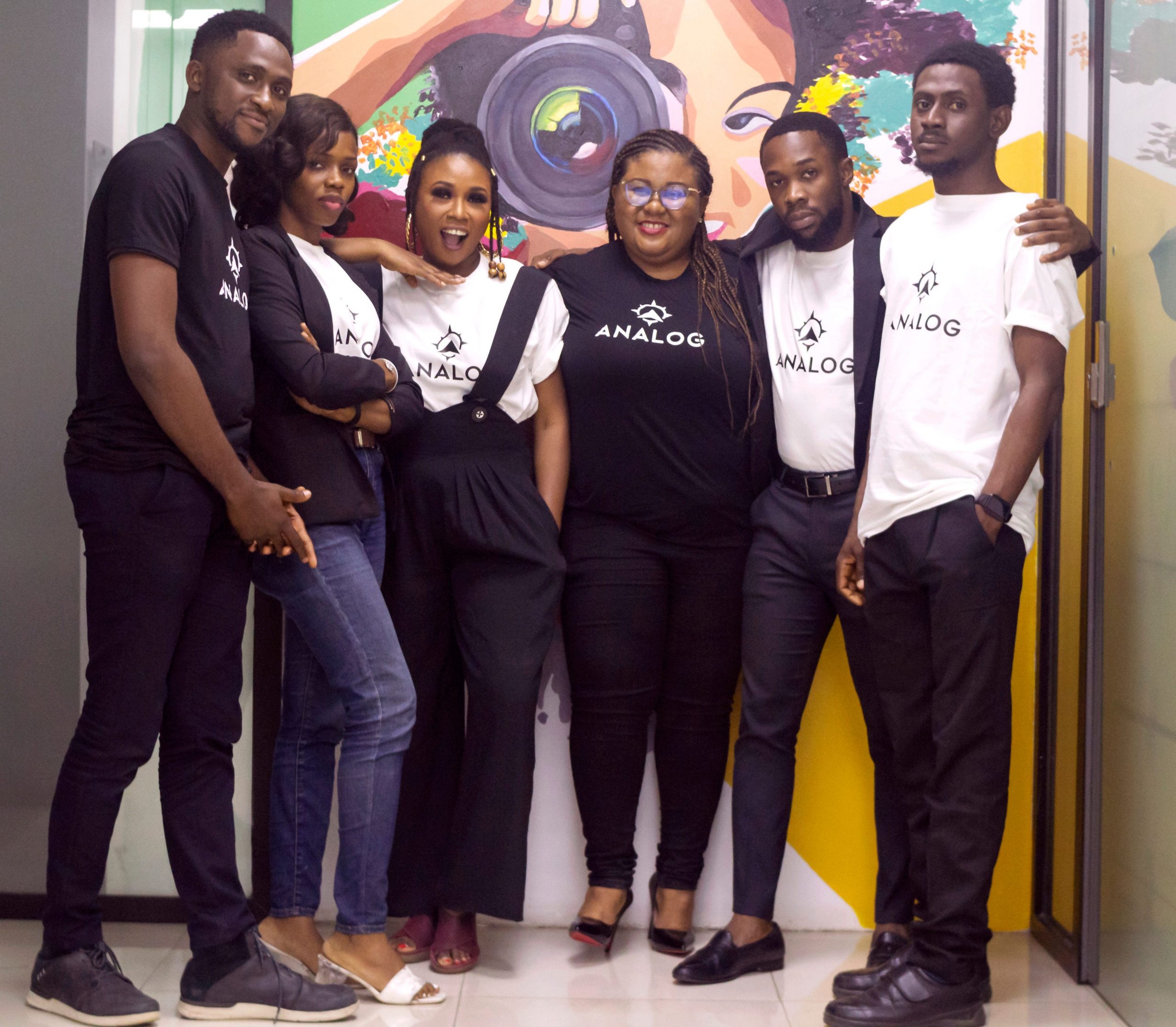 Six Black people stand wearing black and white clothing looking at the camera smiling