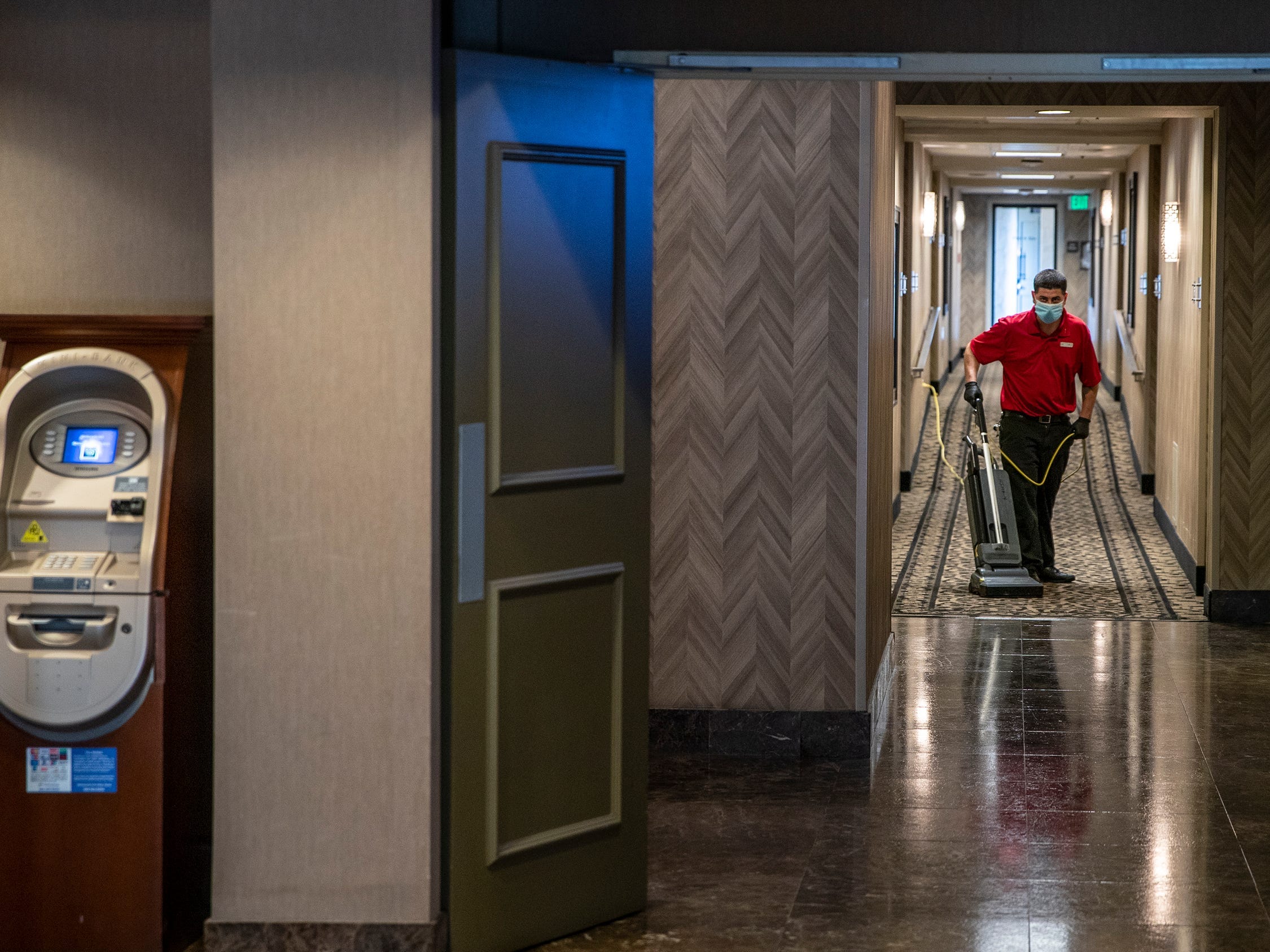 A man wearing a mask vacuums a hotel hallway.