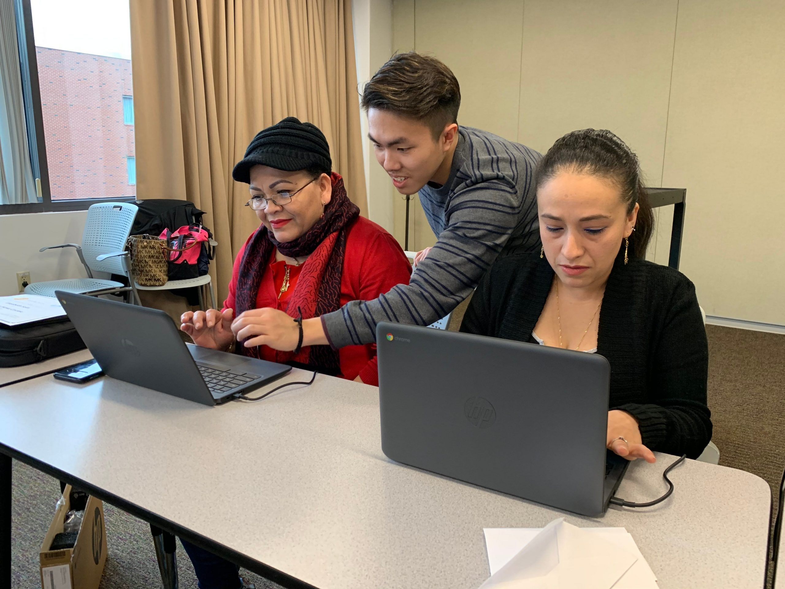 two women work on laptops while being assisted by a man behind them