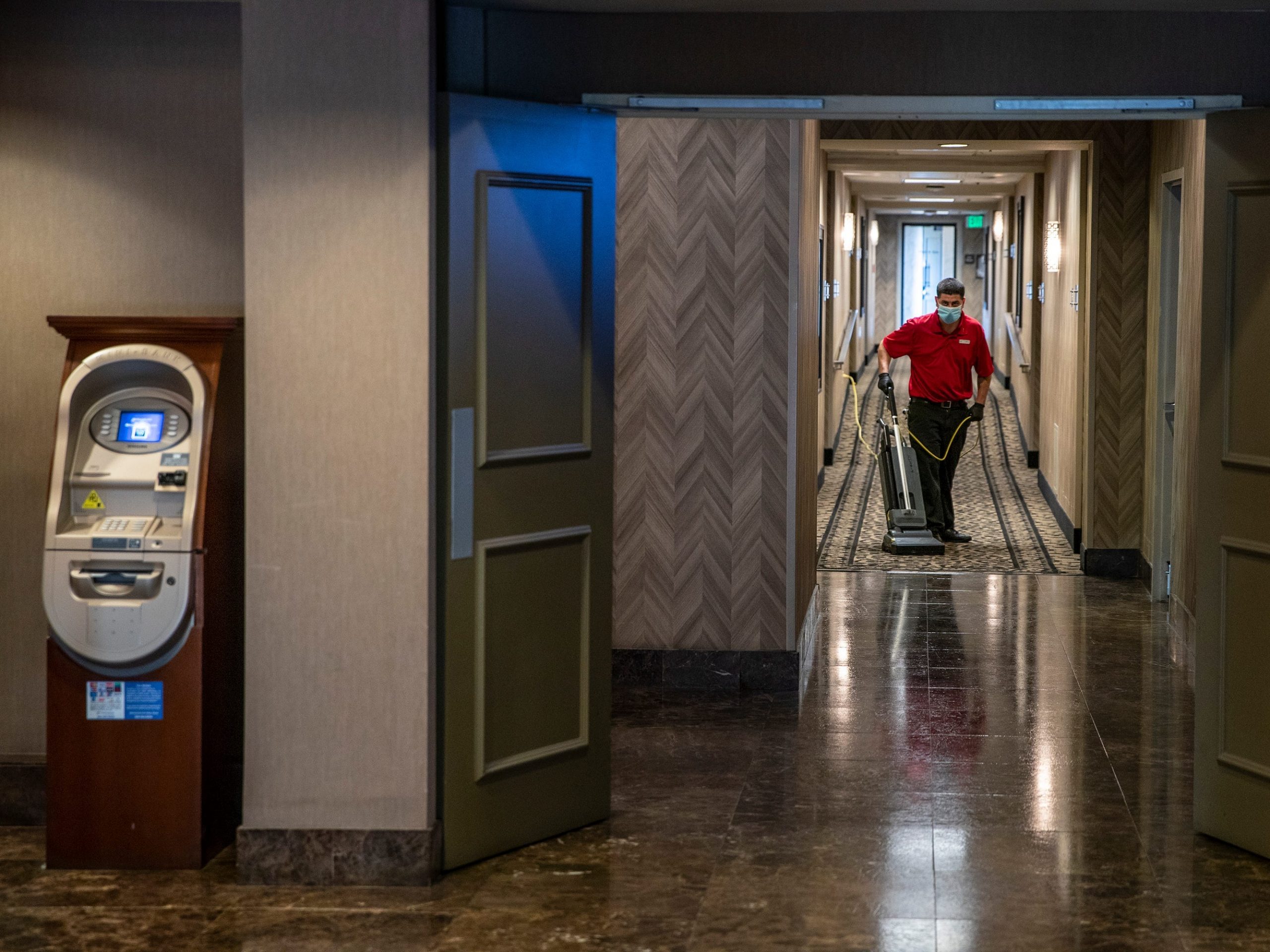 A man wearing a mask vacuums a hotel hallway.