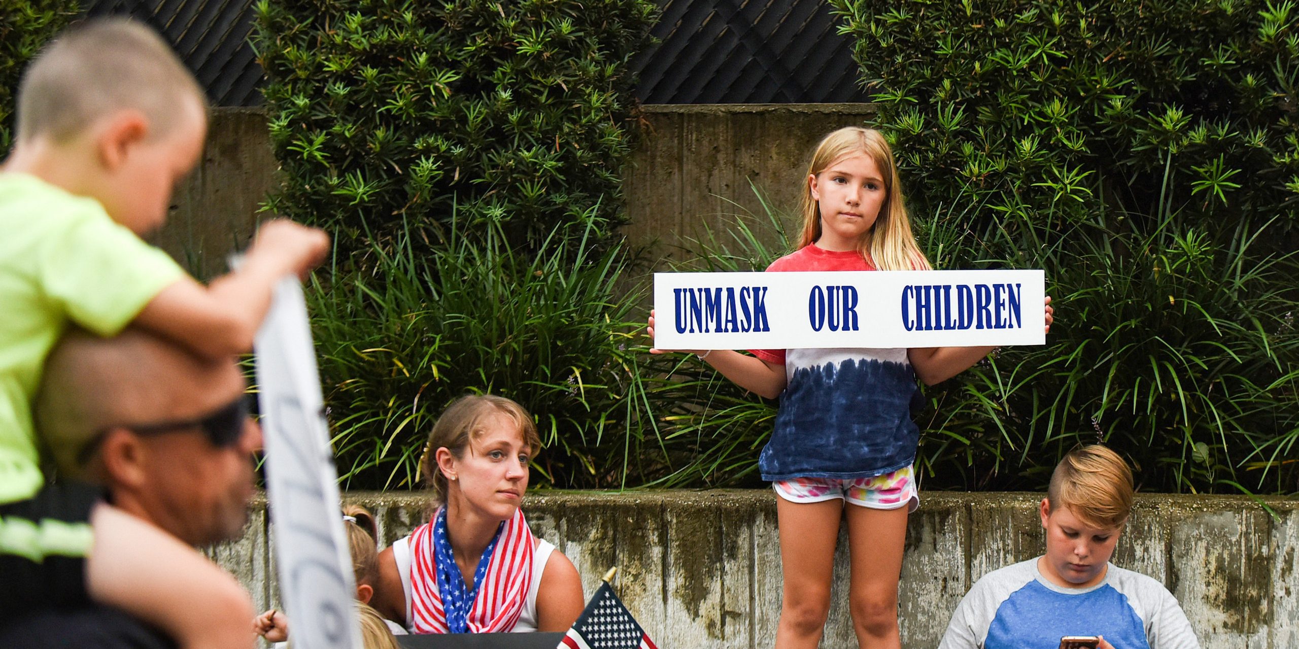 A child holds a sign that reads "Unmask our Children."