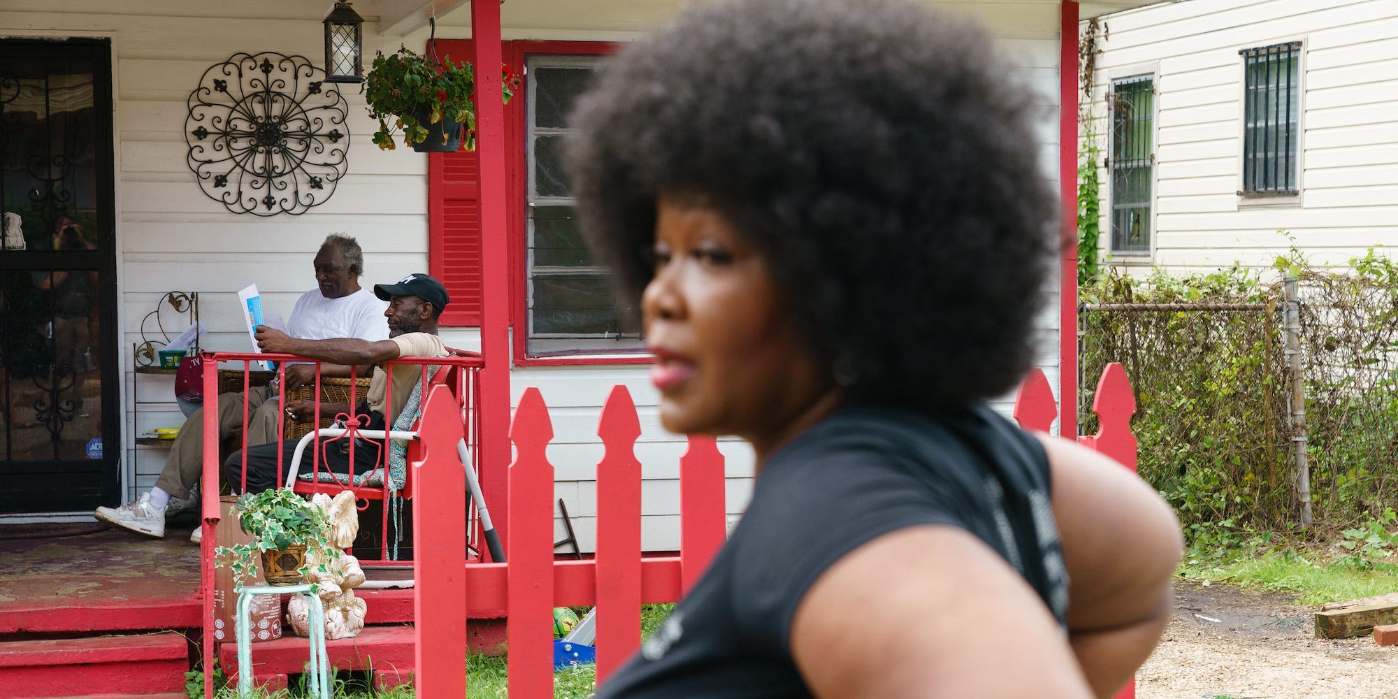 County Commissioner Sheila Tyson stands in the foreground in front of a house with a red-painted porch and picket fence. Two Black residents sit on the porch in the background.
