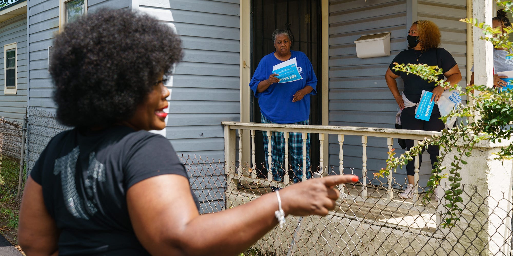 Jefferson County Commissioner Sheila Tyson, wearing black and pointing, stands in the foreground while two Black people on a porch hold papers in the background, during Tyson's vaccine outreach work.