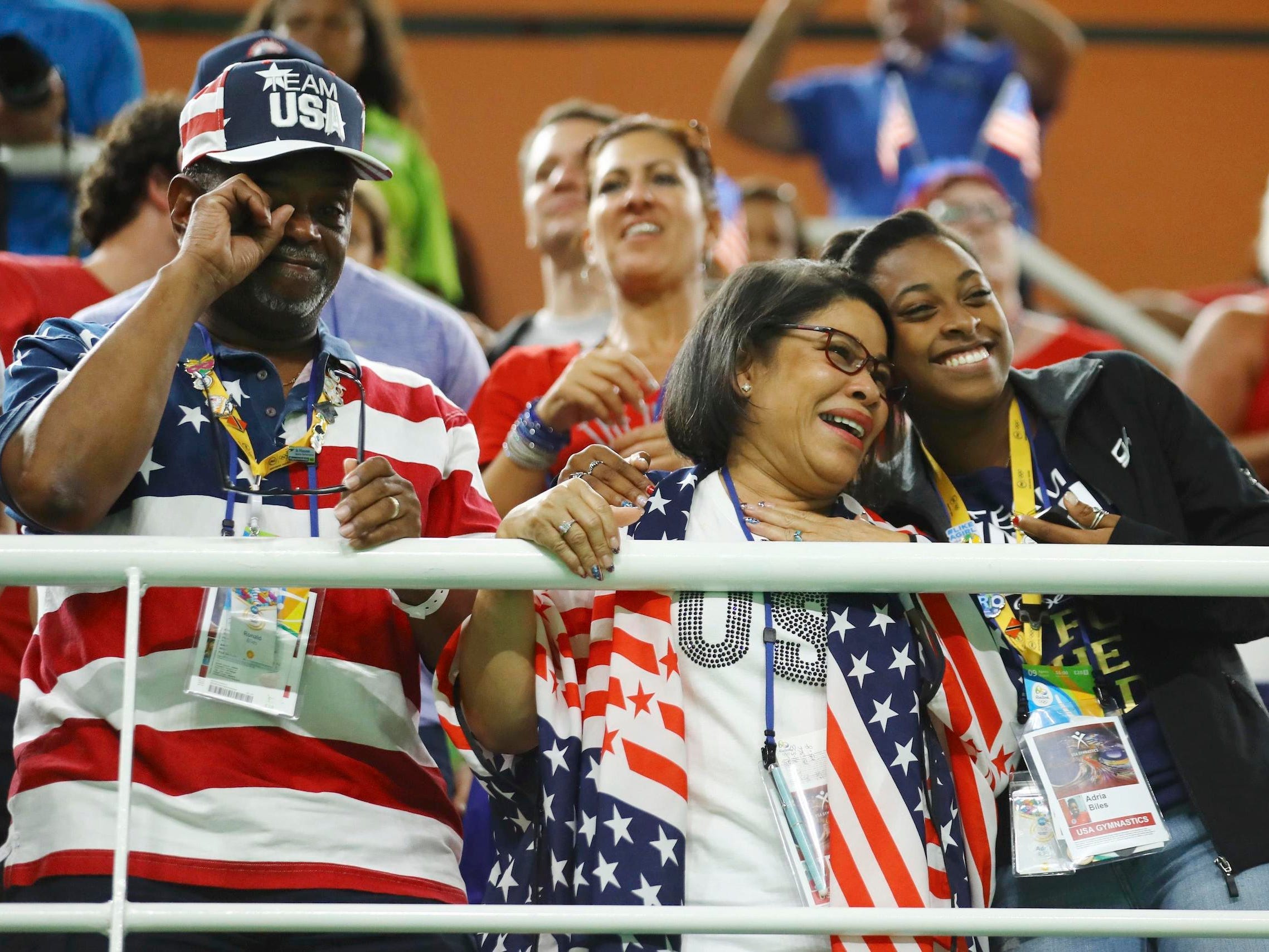 Ron, Nellie, and Adria Biles, wearing USA hats and shirts, watch Simone Biles compete from the stands.