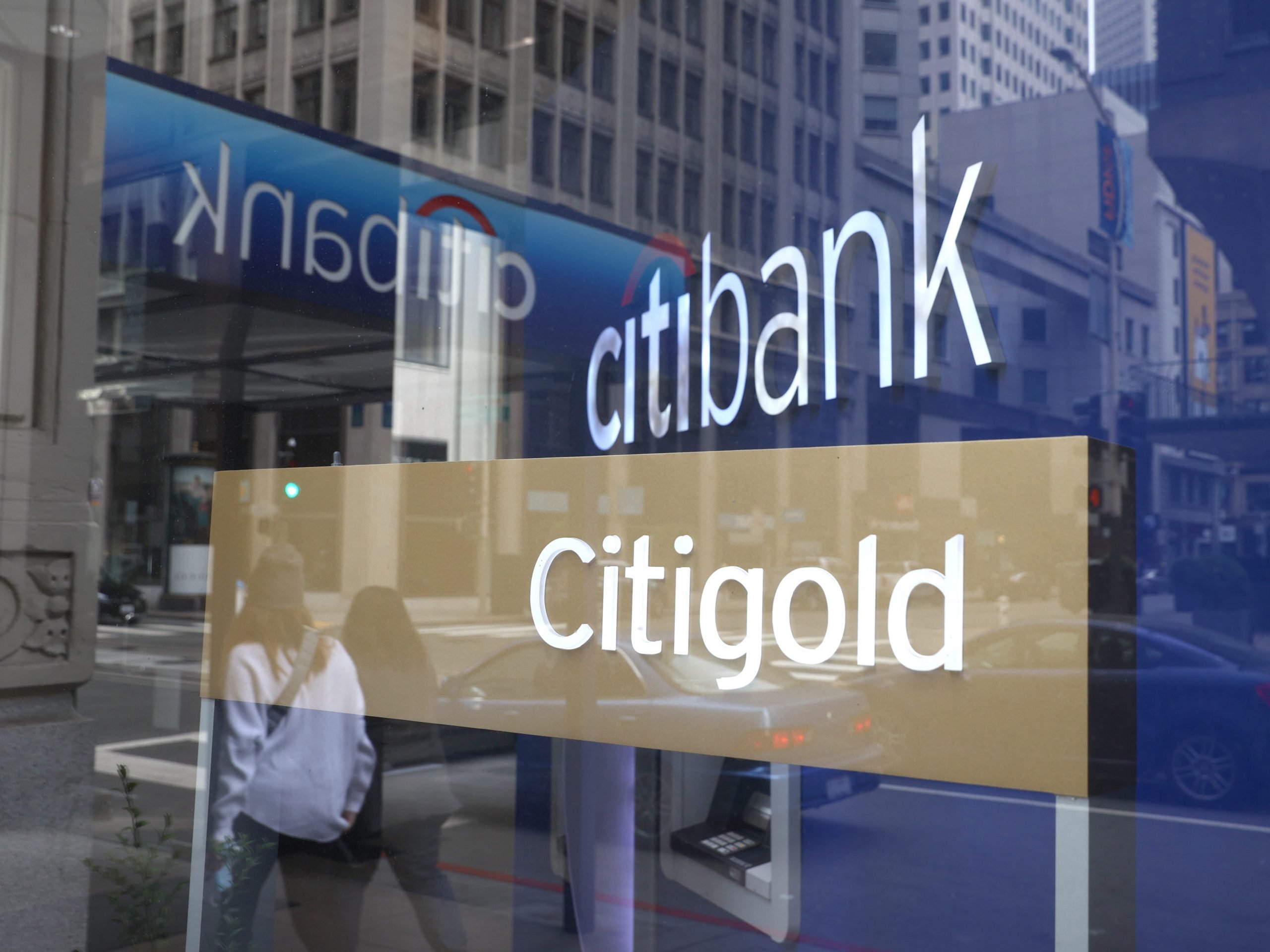 Two blue signs and a golden sign, all with the Citi logo, are seen reflected in front of a bank branch in San Francisco.