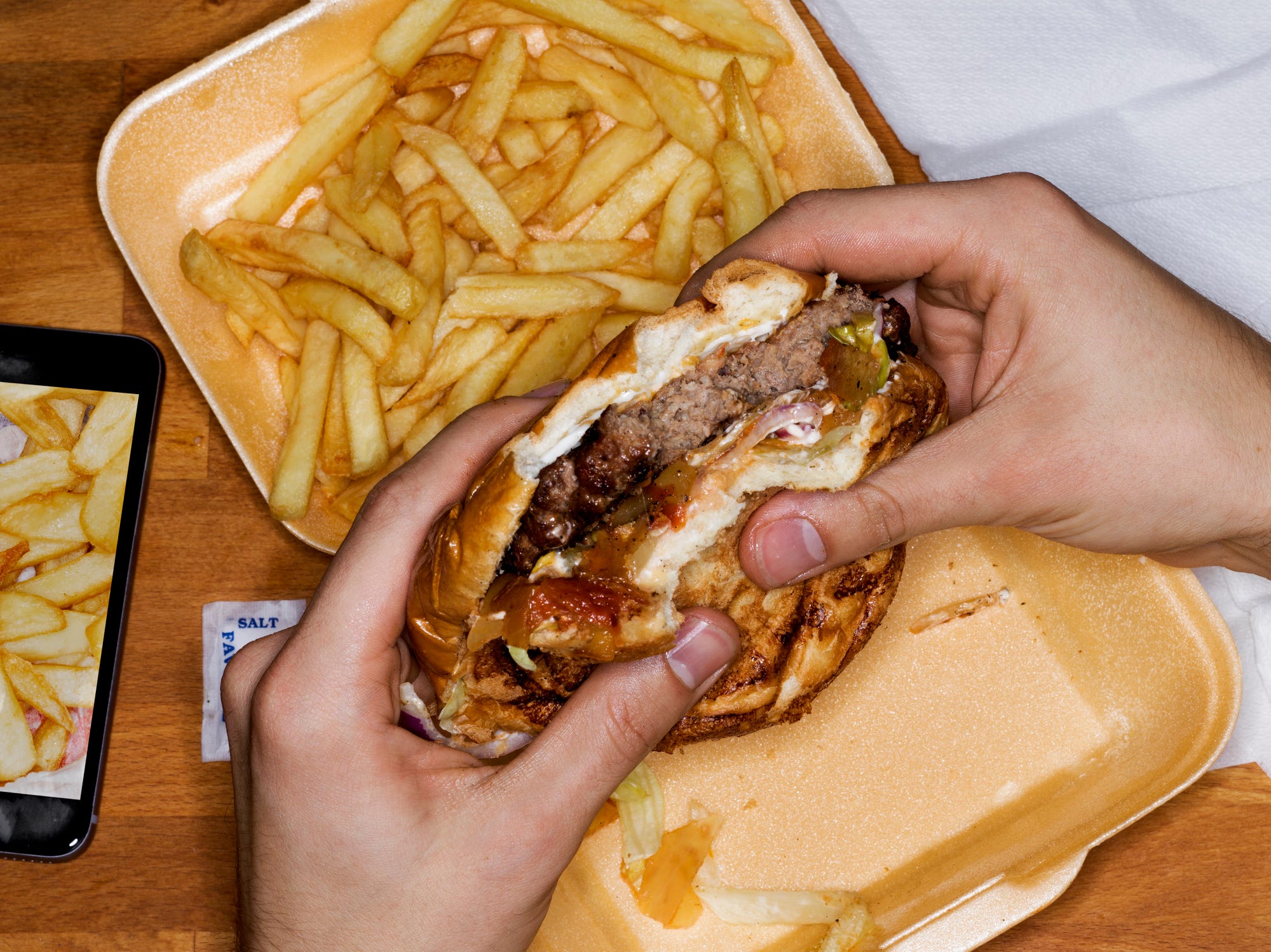 A pair of hands holding a half-eaten hamburger over a takeout container full of french fries