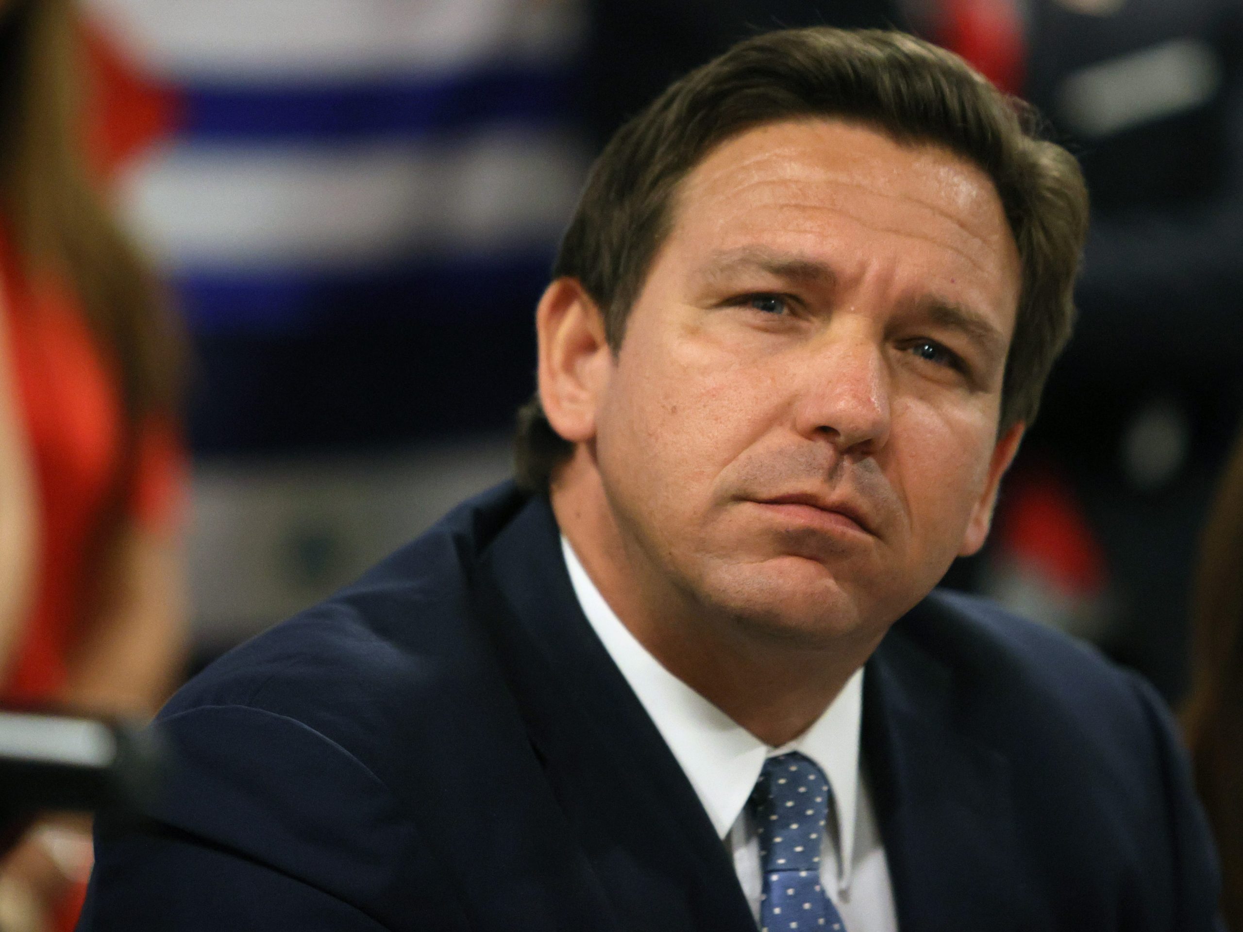 Florida Governor Ron DeSantis wears a black suit and blue tie and looks into the distance.