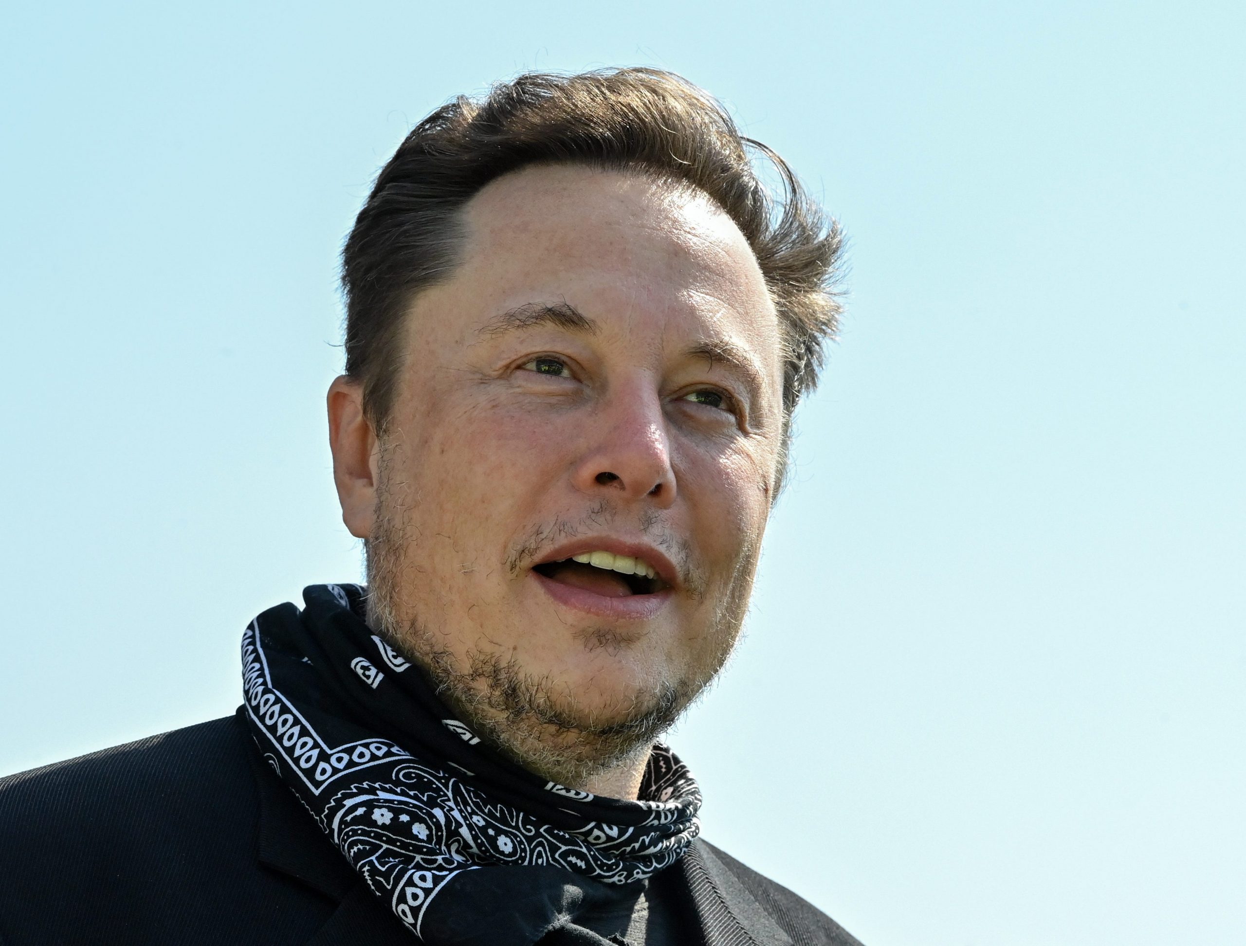 Tesla CEO Elon Musk wears a black and white bandana around his neck in front a light blue sky.