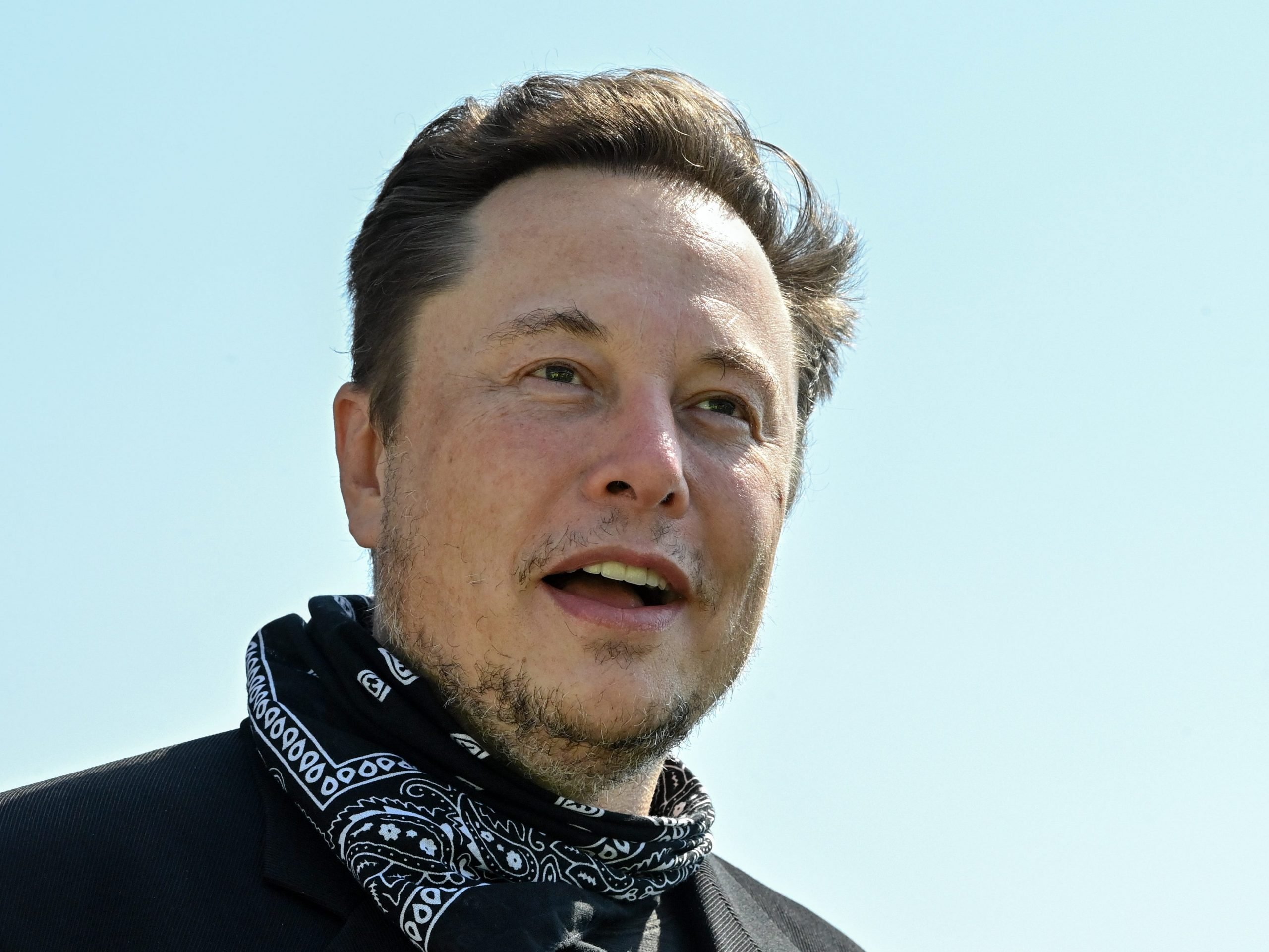 Tesla CEO Elon Musk wears a black and white bandana around his neck in front a light blue sky.