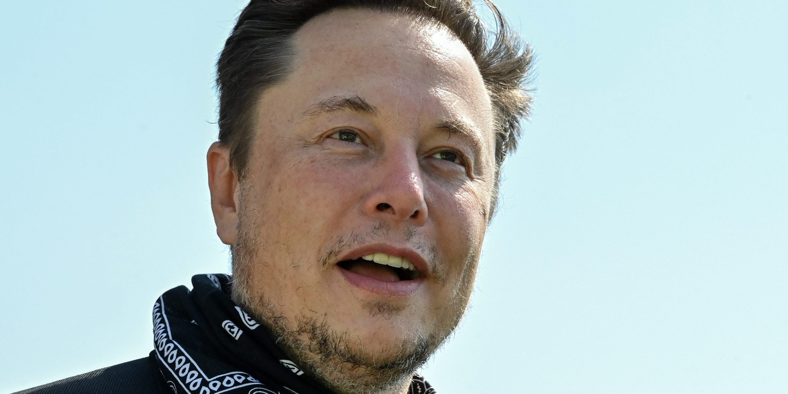 Tesla CEO Elon Musk wears a black and white bandana around his neck in front a light blue sky.