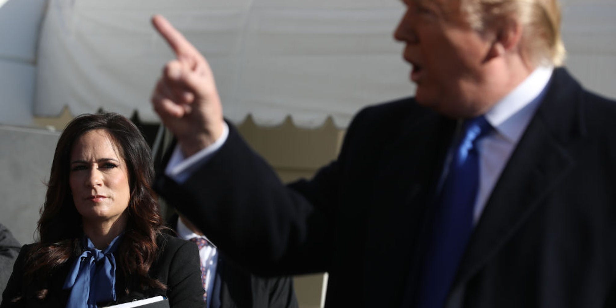 Stephanie Grisham stands behind Donald Trump, who is speaking with his finger held in the air.