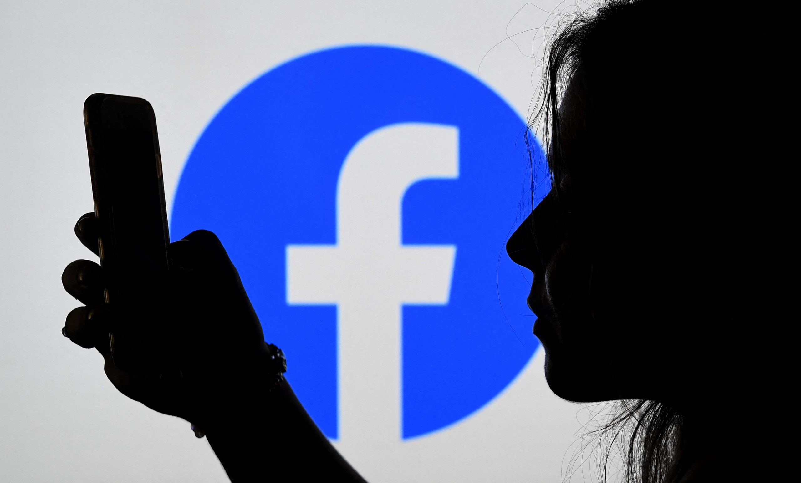 A person looks at. a smart phone with a Facebook logo displayed in the background