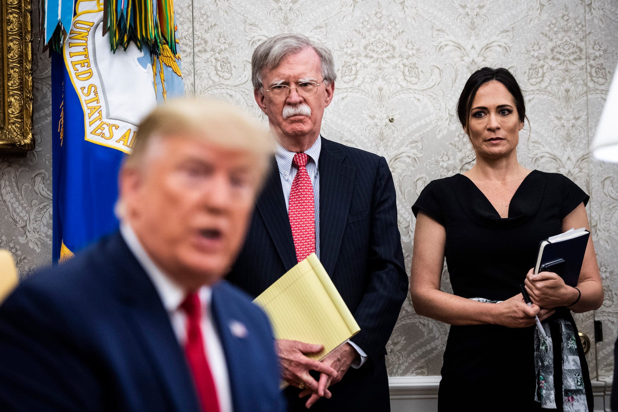 Stephanie Grisham and John Bolton looking at Donald Trump.