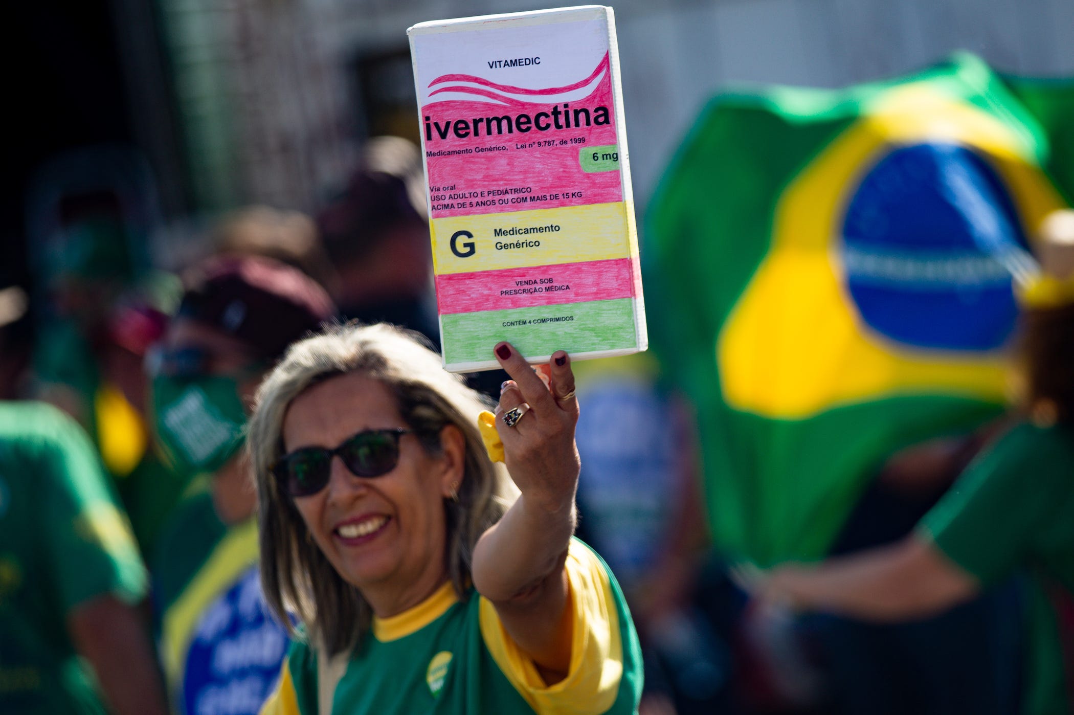 a woman wearing the colors of Brazil's flag and smiling holds up a giant box of ivermectin