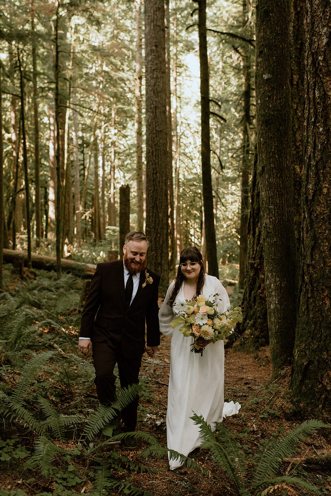 Two people, one in a black suit and one in a white wedding dress, walking through the forest together