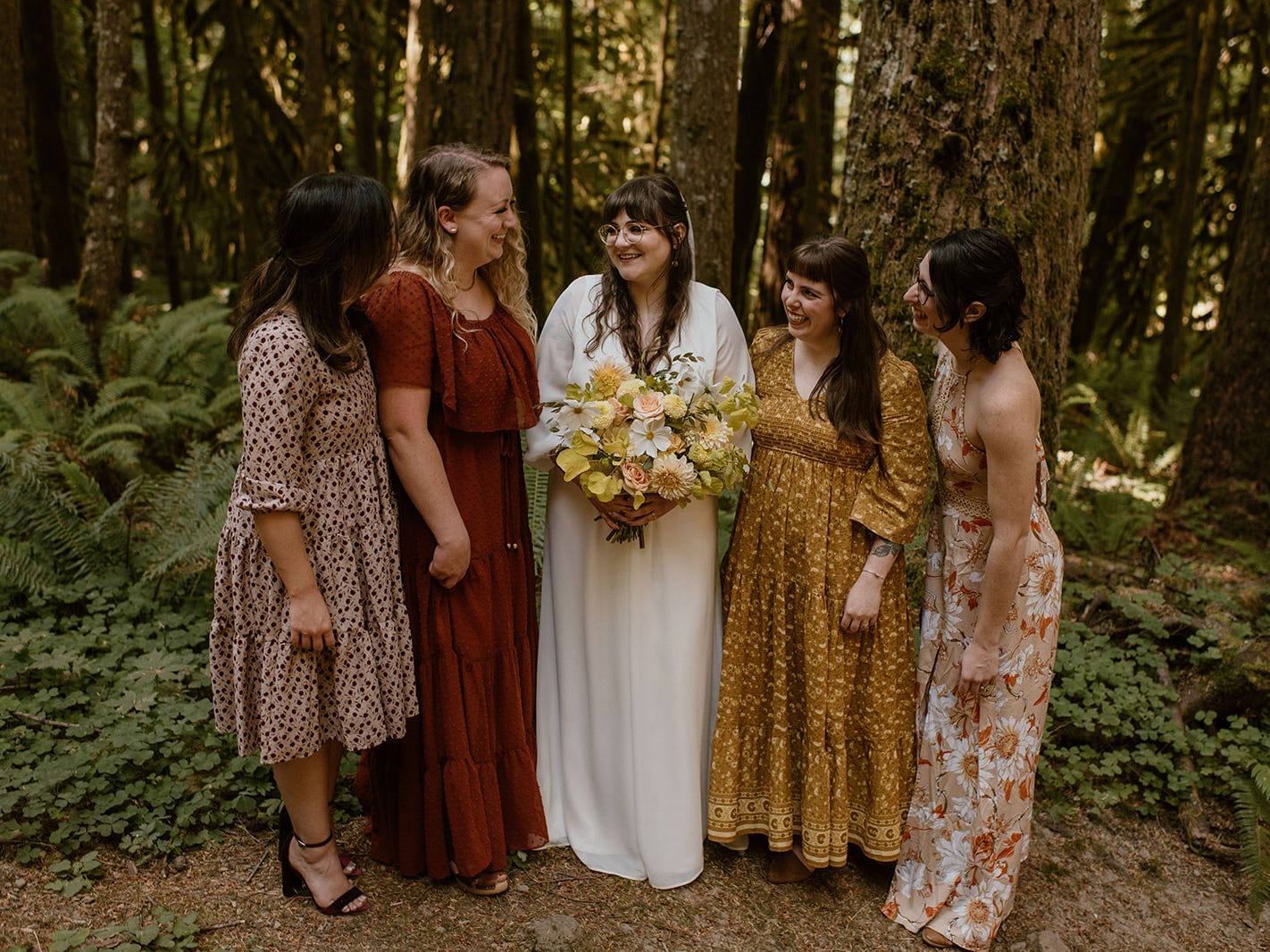 A person in a wedding dress holding a bouquet surrounded by bridesmaids wearing fall tones, in a forest