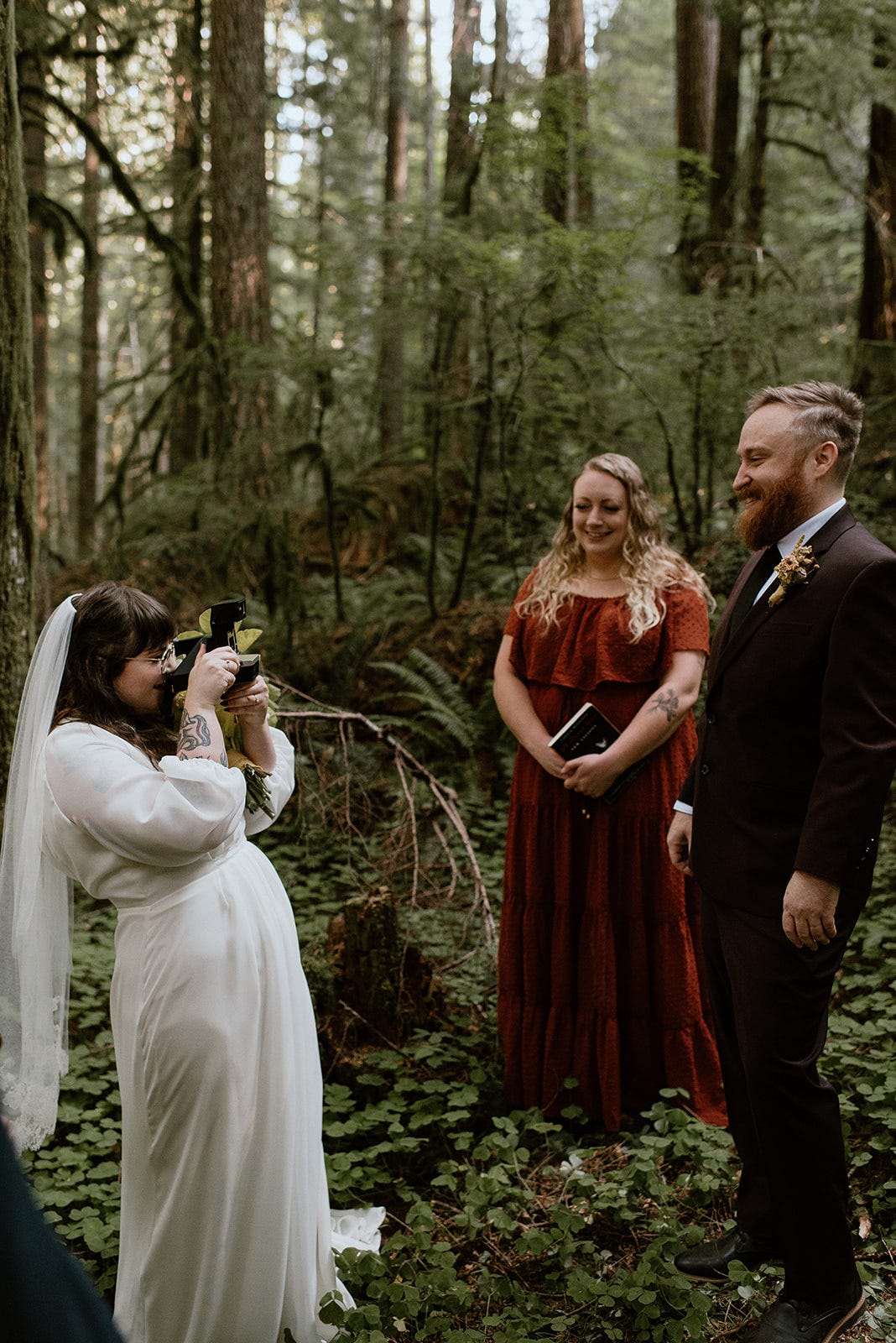 A person in a wedding dress taking a picture of a person in a black suit in the forest