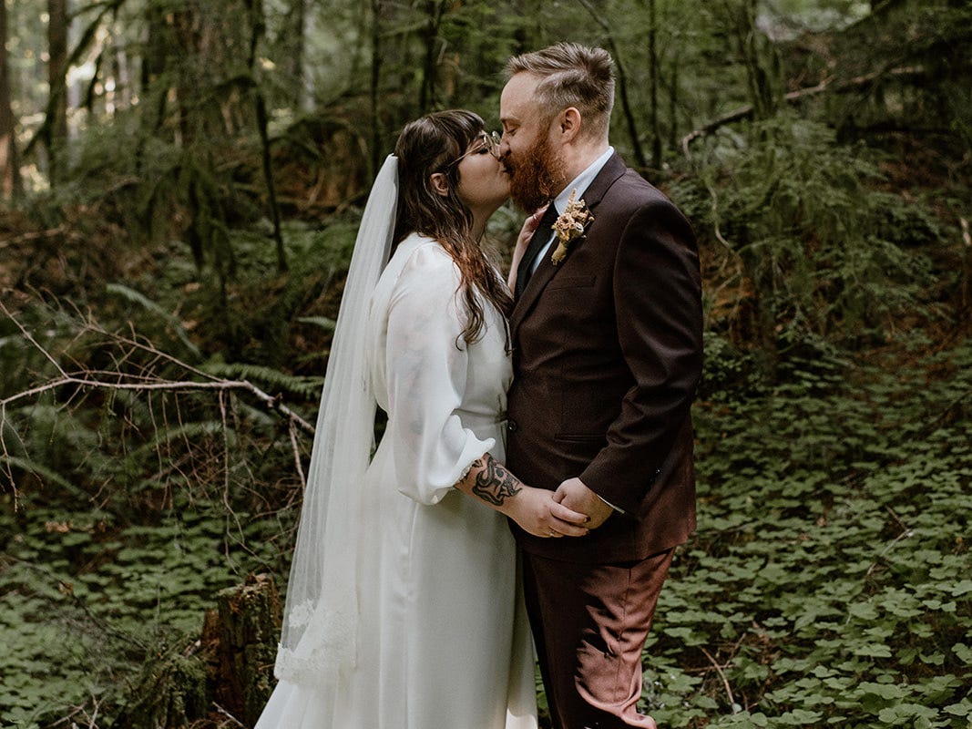 A couple getting married, one person wearing and wedding dress and the other wearing a black suit, kissing in a forest