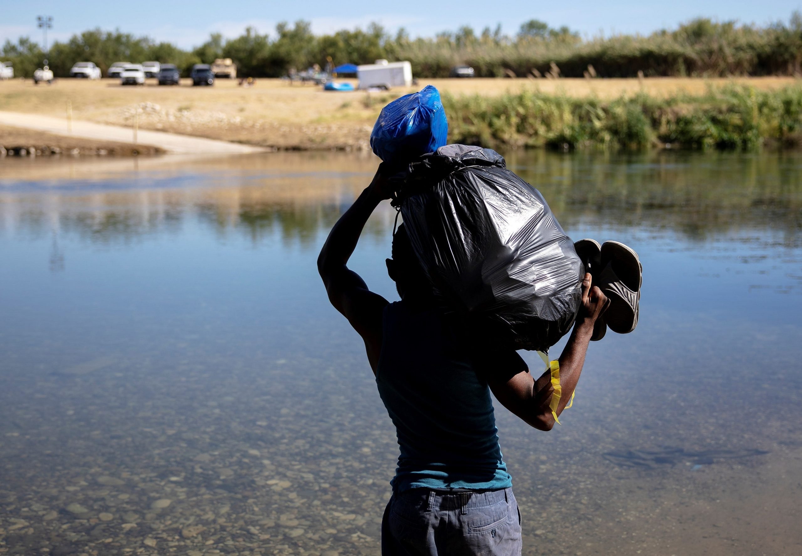 Haitian migrant, on US-Mexico border, seen from behind attempting to cross a river carrying a trash bag of belongings atop their shoulder.