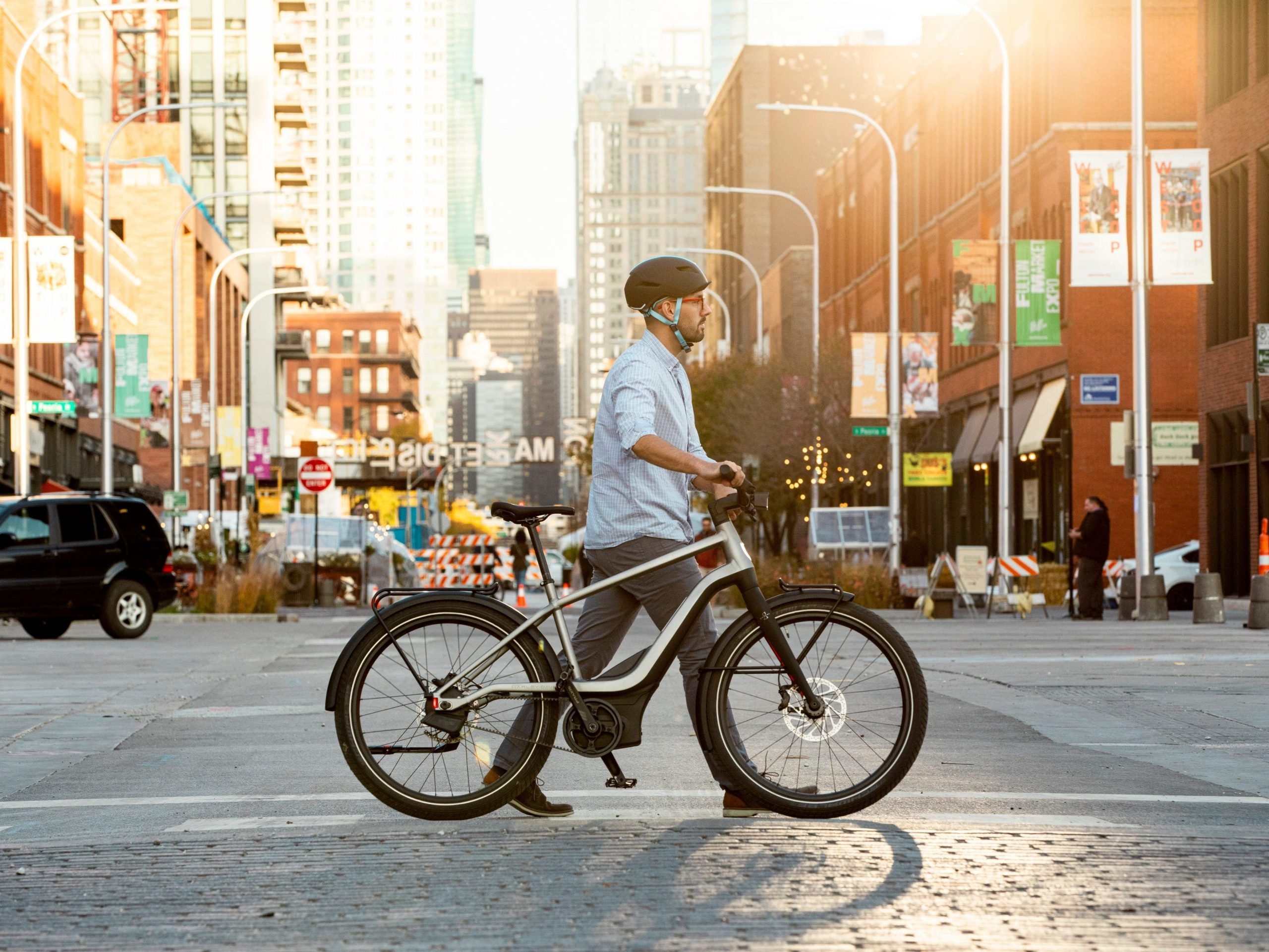 A man walks a Serial 1 electric bike across a street. He is wearing a helmet, blue shirt, and dress pants.