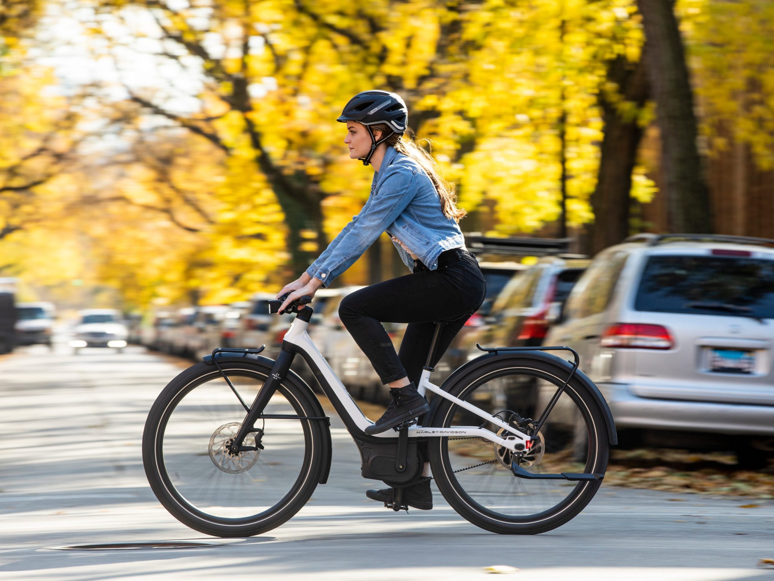 A woman wearing a bike helmet, jean jacket, black pants and boots rides a Serial 1 electric bike on a street.