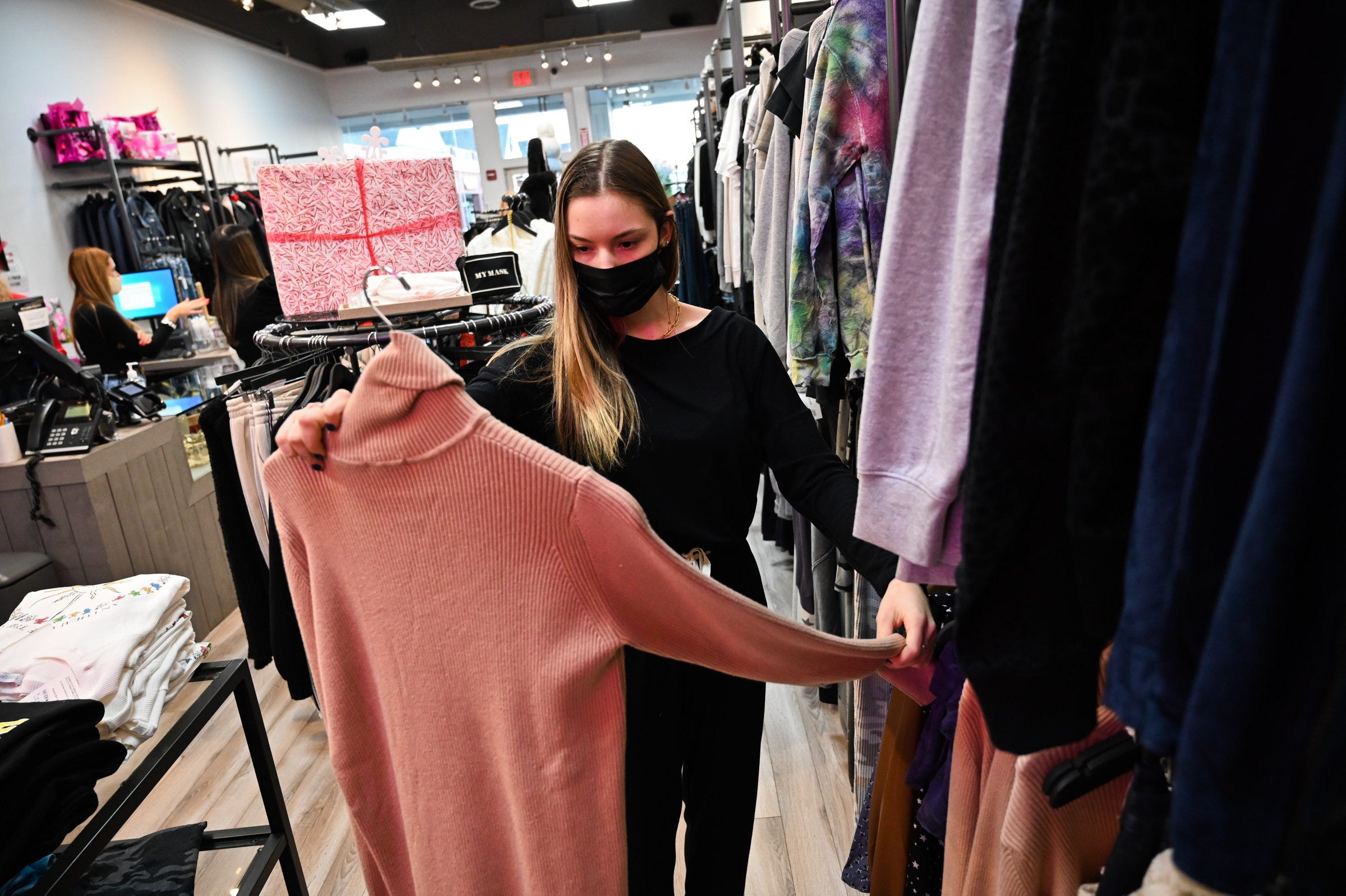 Woman wearing face mask holds up pink turtleneck sweater while clothes shopping