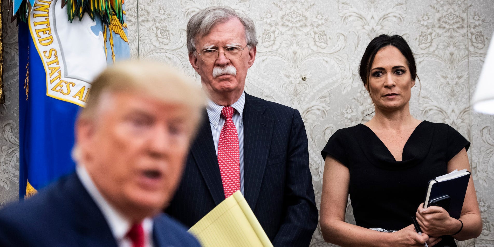 Stephanie Grisham and John Bolton looking at Donald Trump.