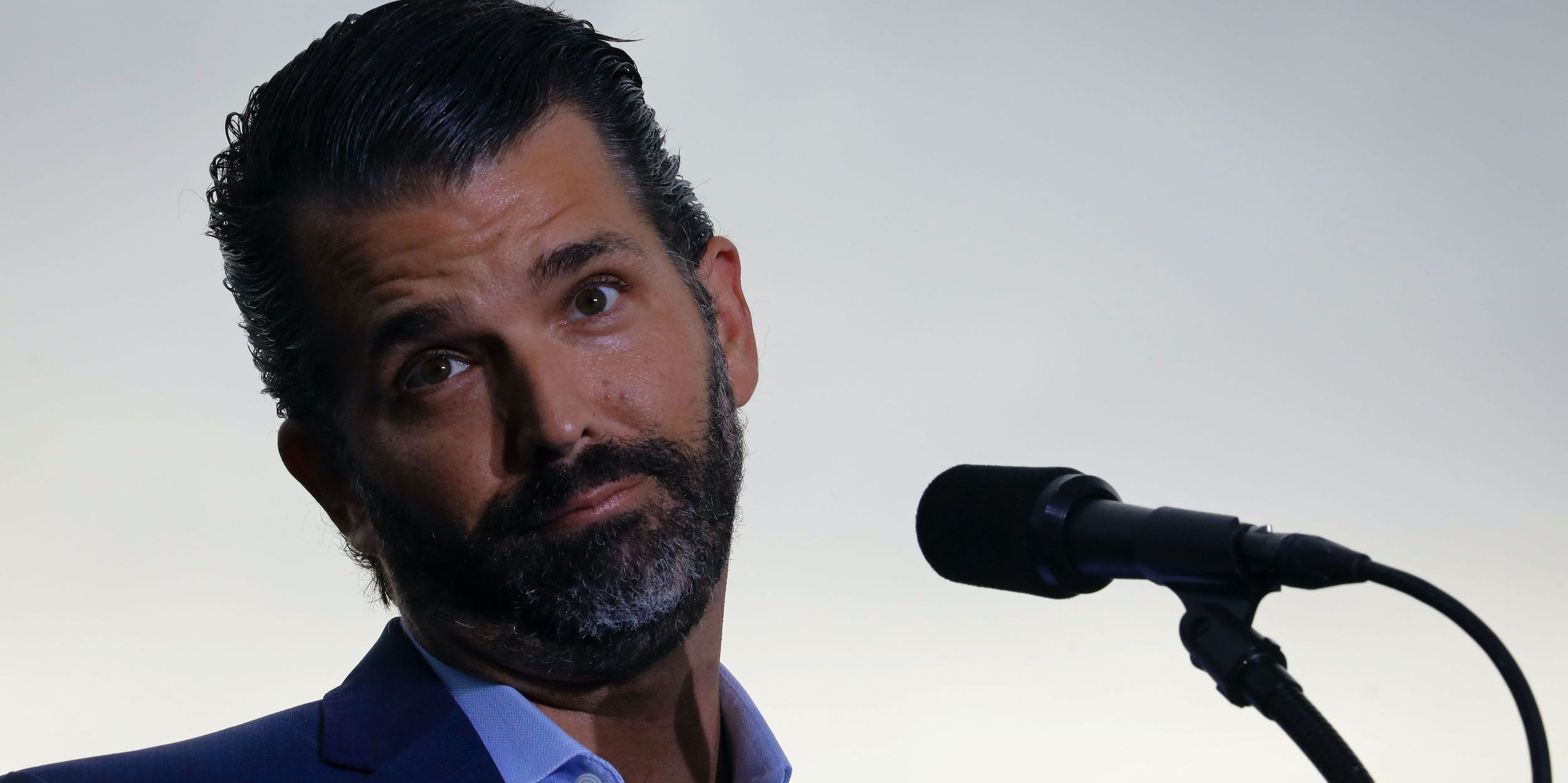 Donald Trump Jr. tilts his head and raises his eyebrows with his lips pursed behind the mic at a Trump rally.