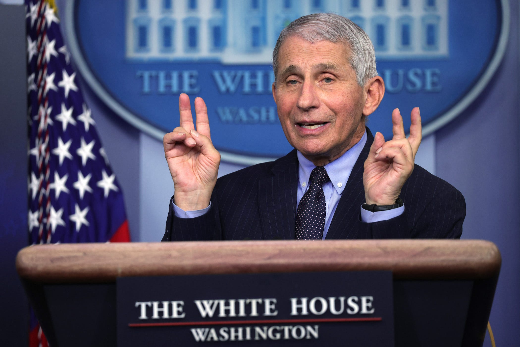 fauci in a suit at the podium of the white house press briefing room, using air quotes