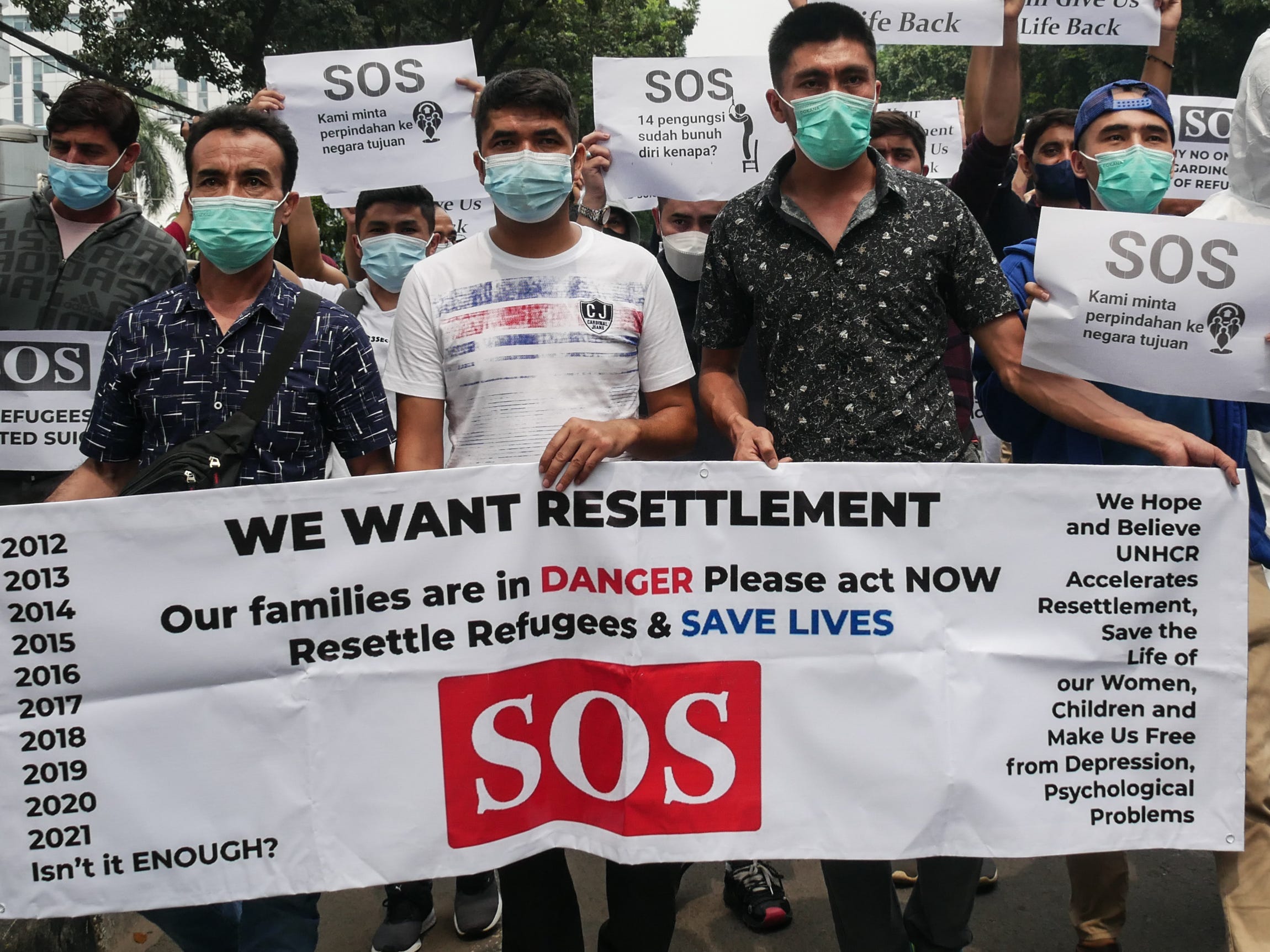 A crowd of Afghan people with masks on hold a sign that reads: "We want resettlement. Our families are in danger please act now. Resettle refugees and save lives. SOS"