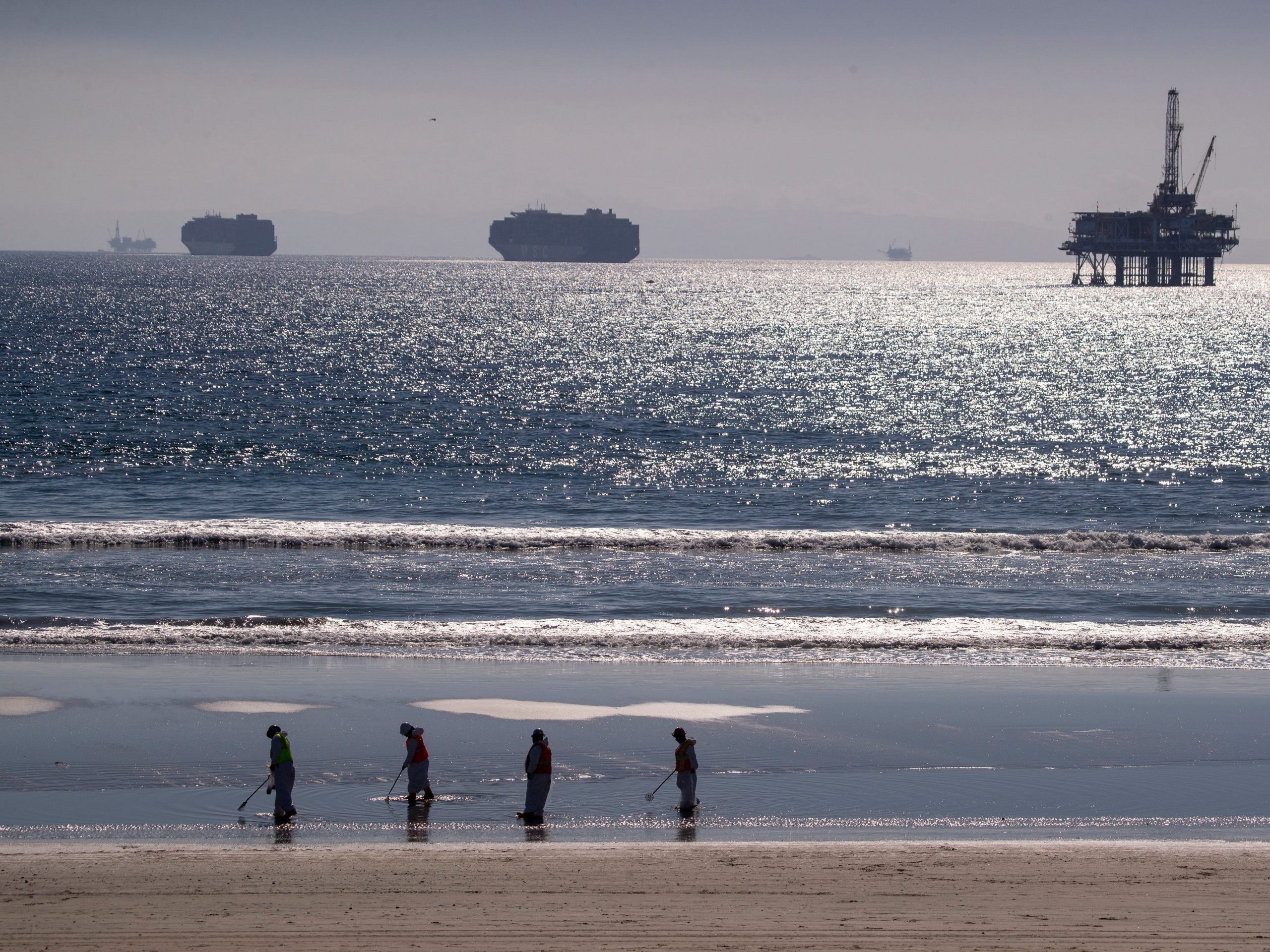 Workers clean a beach with an oil drilling platform in the background.