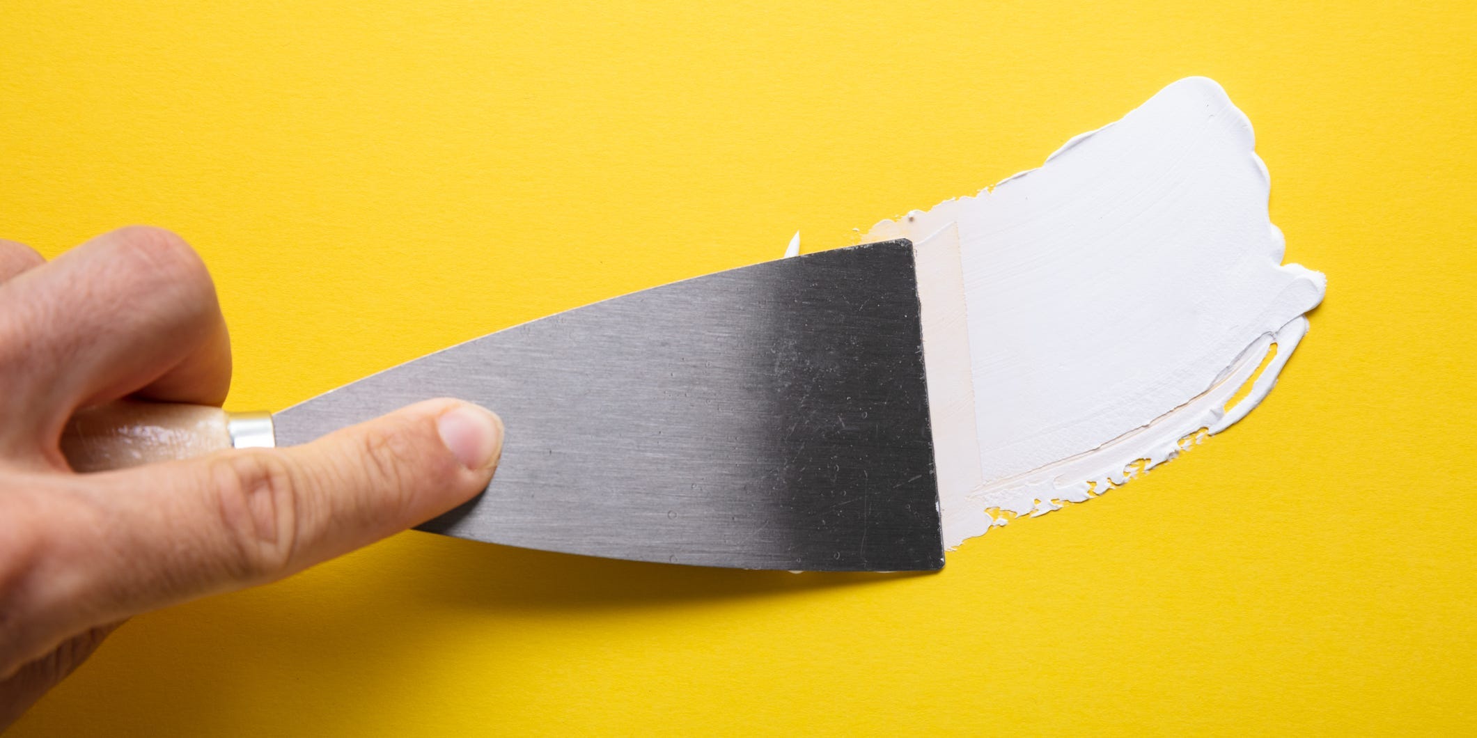 A hand using a putty knife to apply spackle to a yellow wall